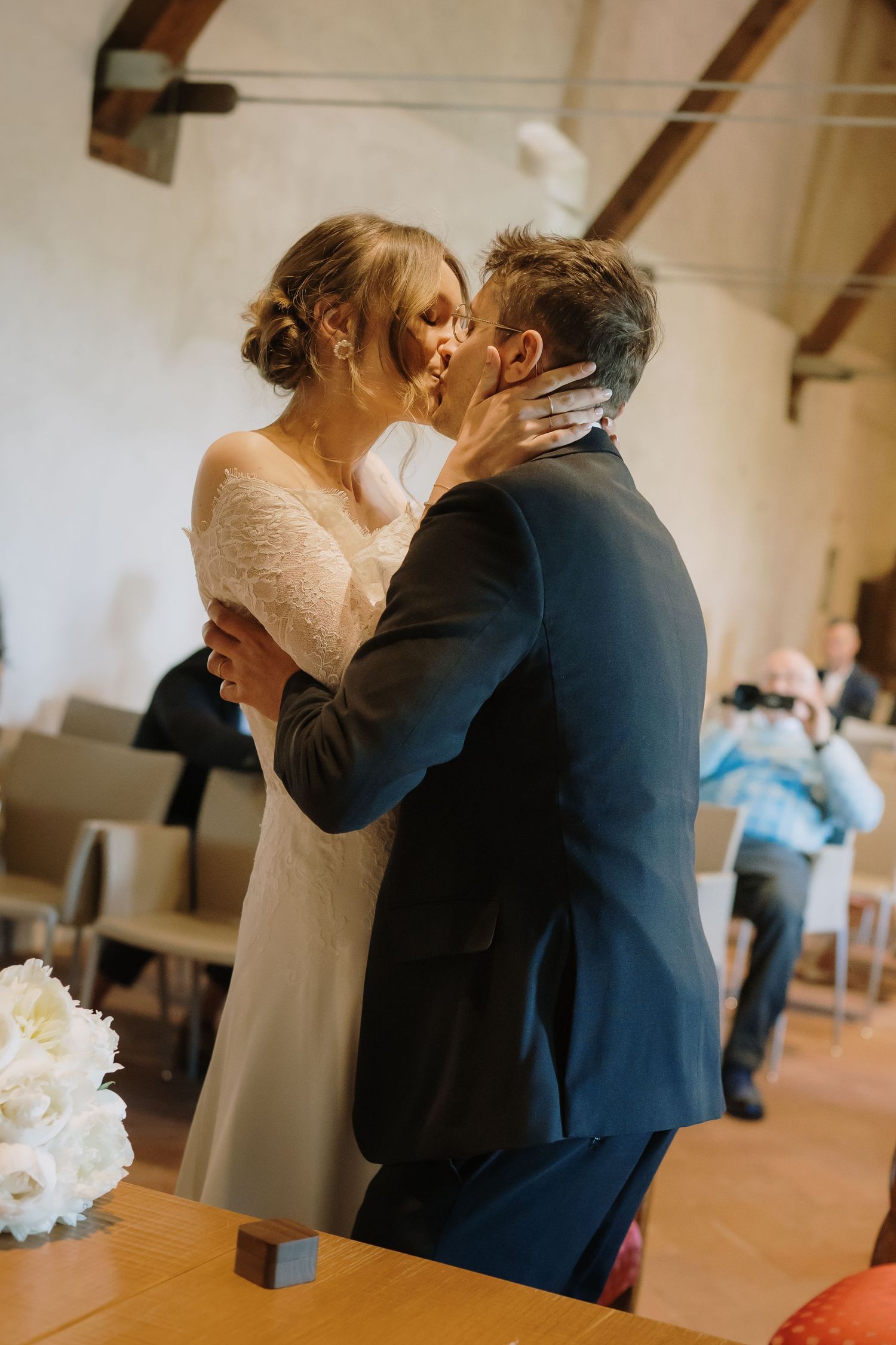 A bride and groom share their first kiss at a wedding ceremony in a rustic venue with wooden beams.