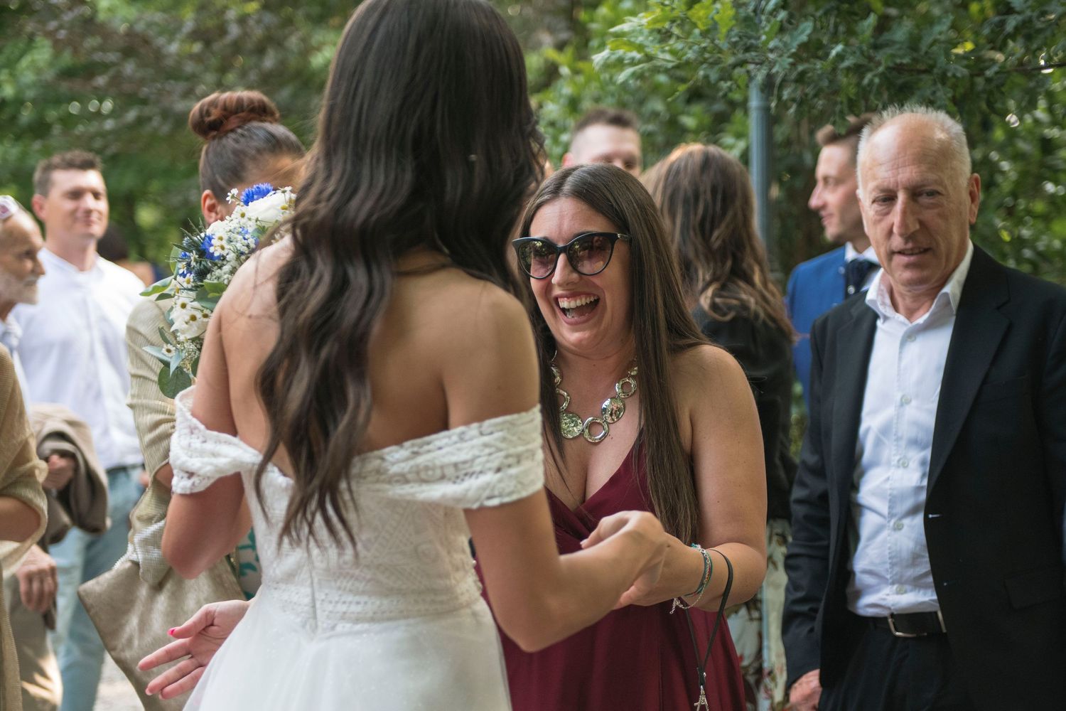 Smiling wedding guest in burgundy dress greets bride in white gown at outdoor reception.