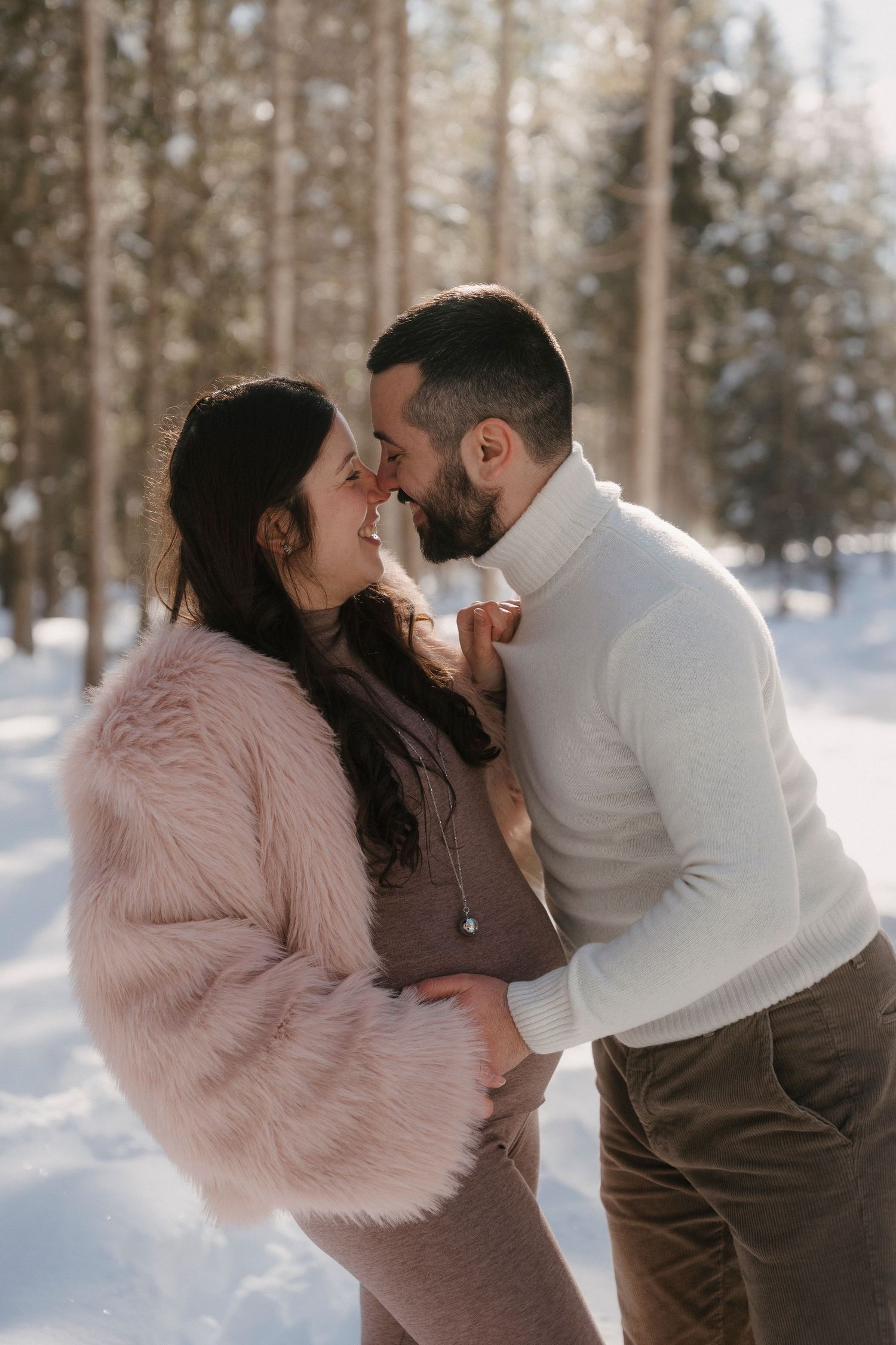 Expecting couple sharing a romantic nose-to-nose moment in a bright snowy winter forest.