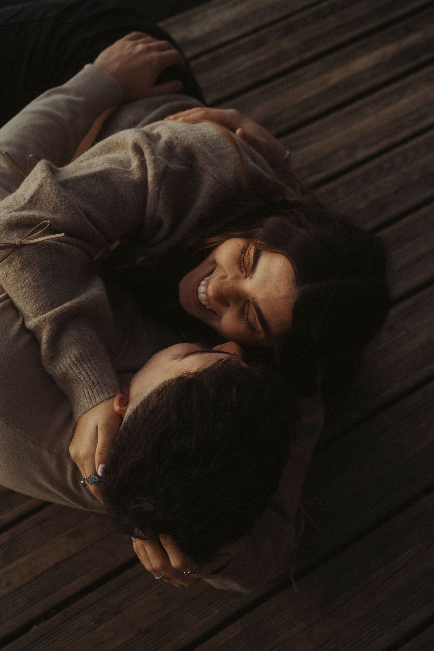 Happy couple lying on a wooden deck, smiling and embracing in warm sunlight.
