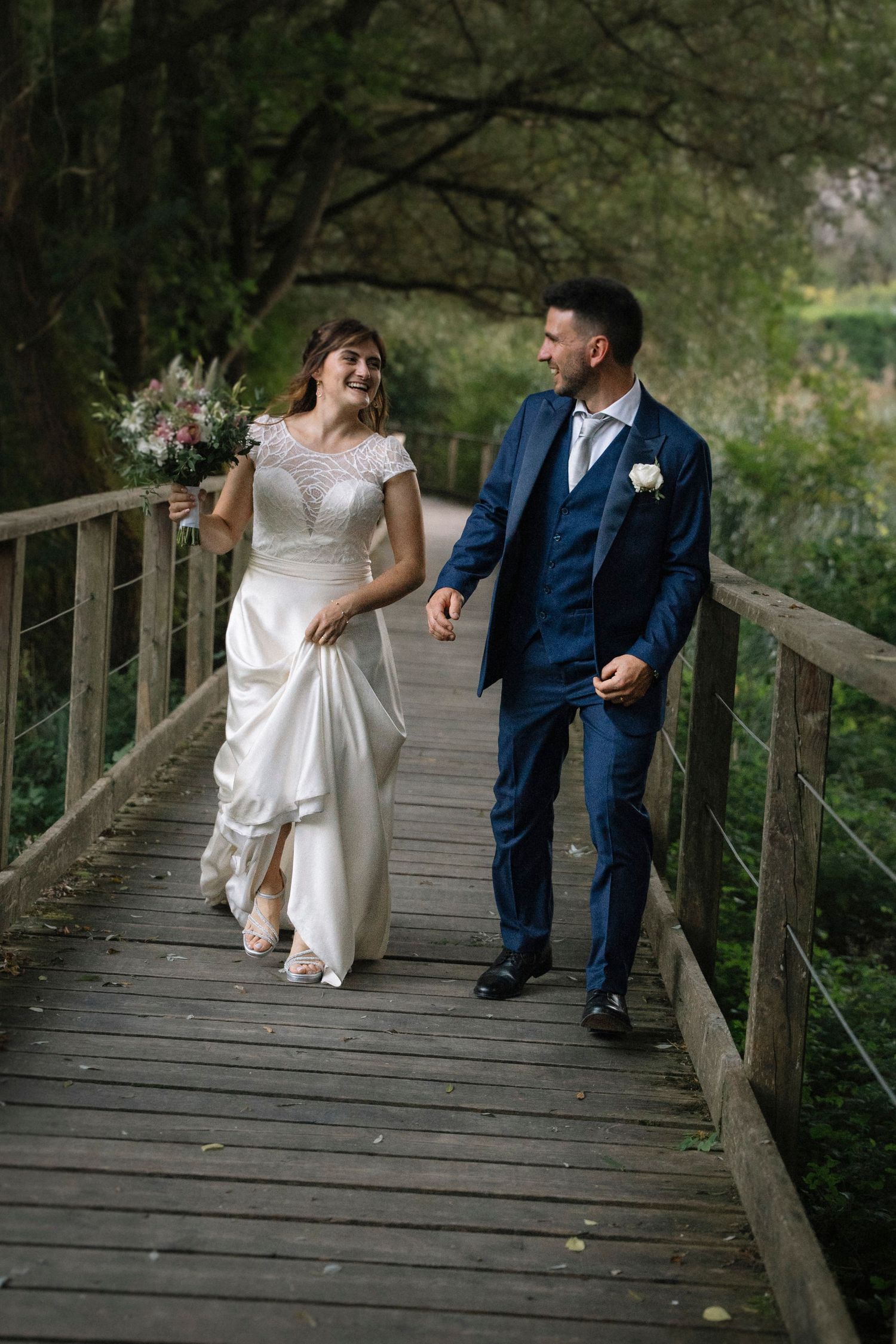 Happy bride and groom walking hand in hand across a wooden bridge surrounded by lush greenery.