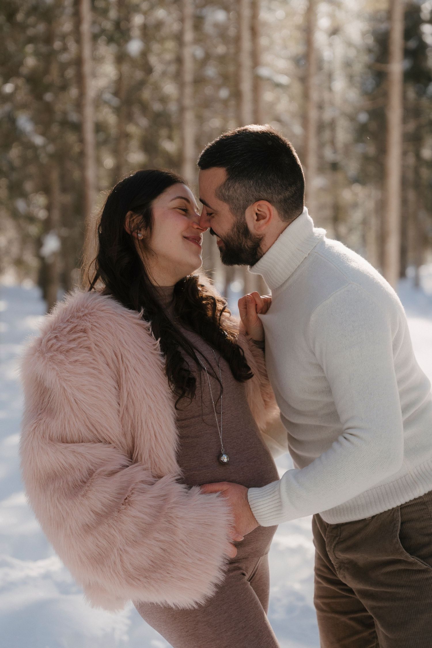 Pregnant couple sharing a tender moment in a snowy winter forest, woman in pink fur coat cradling baby bump.