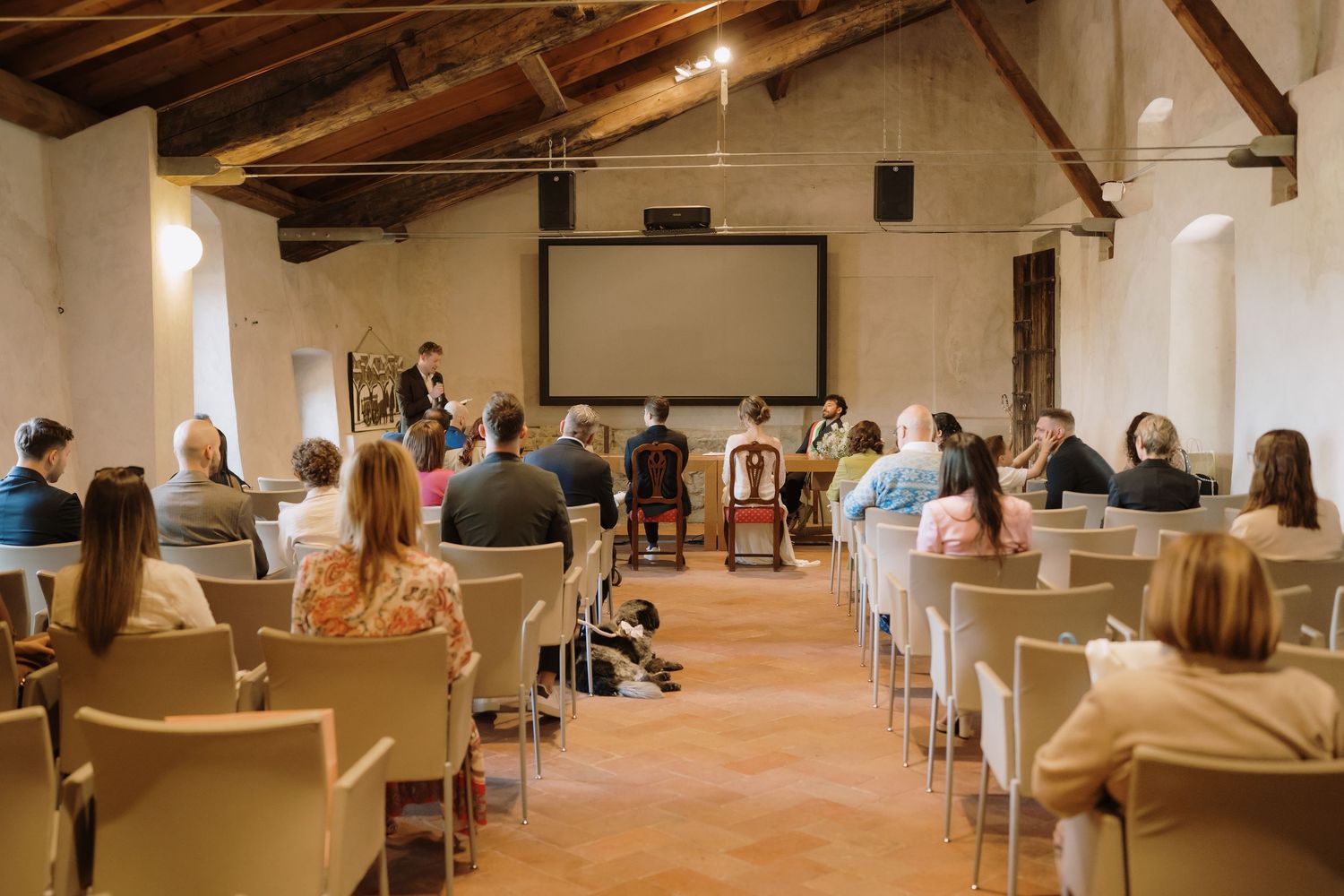 Wedding ceremony in rustic venue with wooden beams, guests seated facing a projection screen and musicians.