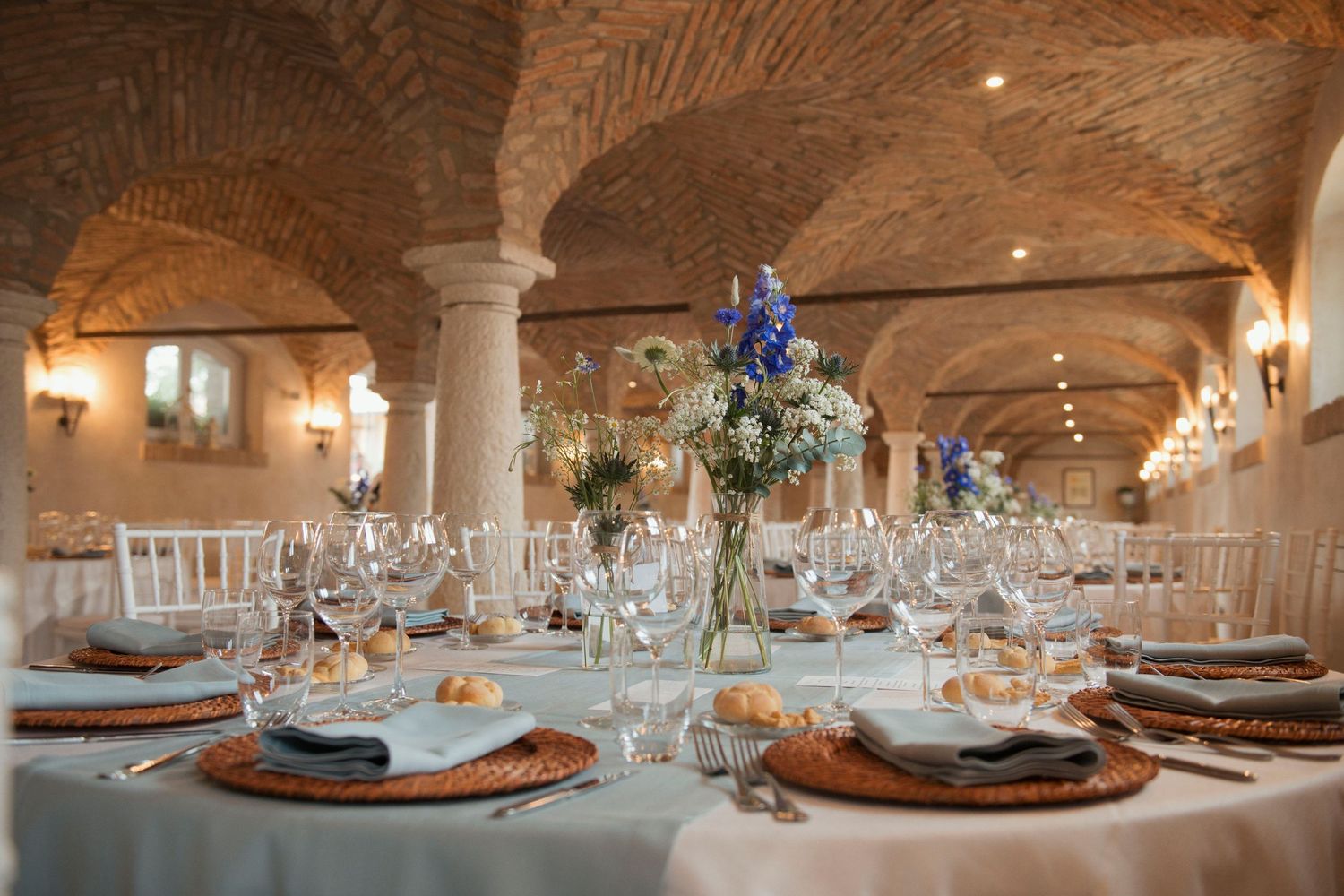 Elegant wedding reception in a rustic stone vaulted hall with floral centerpieces, crystal glassware, and wicker charger plates.