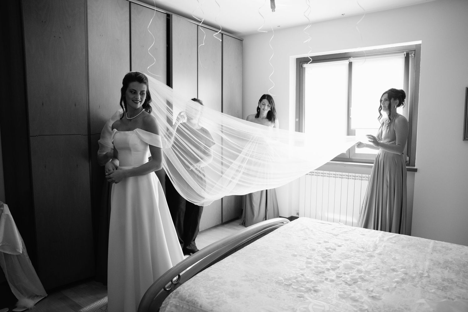 Bride and bridesmaids preparing wedding veil in bedroom, black and white photo.