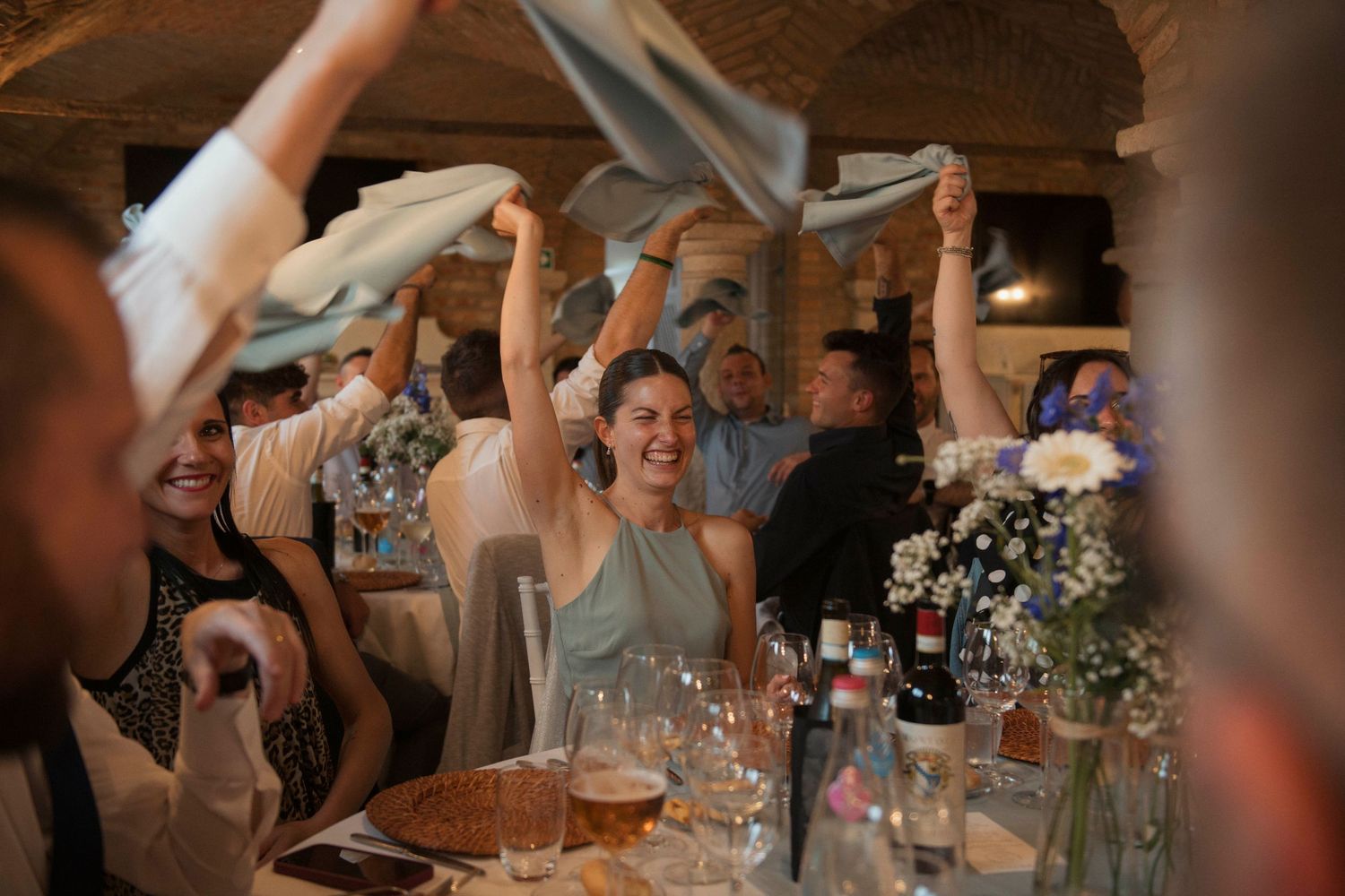 Joyful wedding guests waving napkins in the air during a lively celebration at a rustic reception venue.