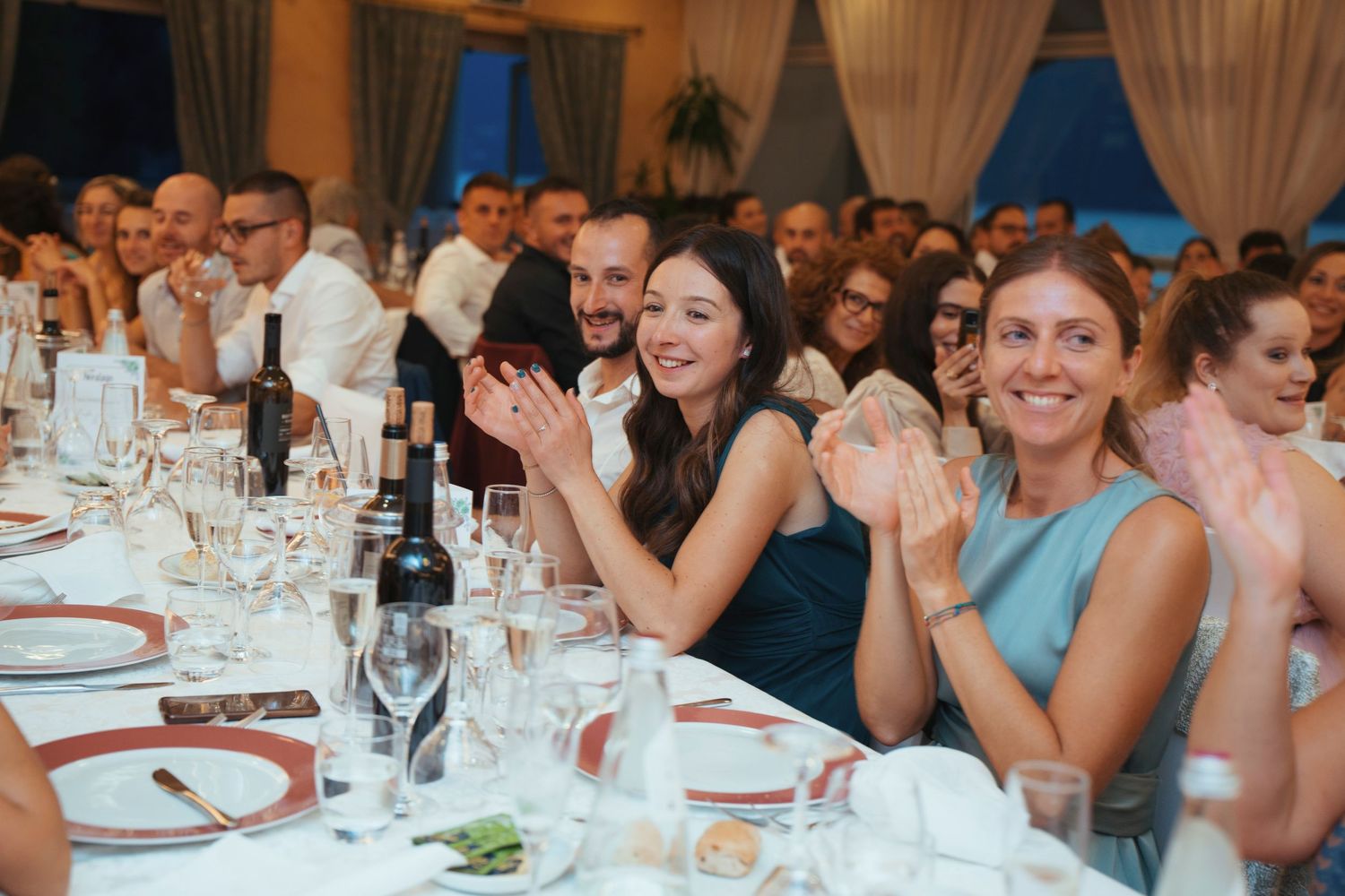 Wedding guests smiling and clapping joyfully at elegantly set tables during a wedding reception.