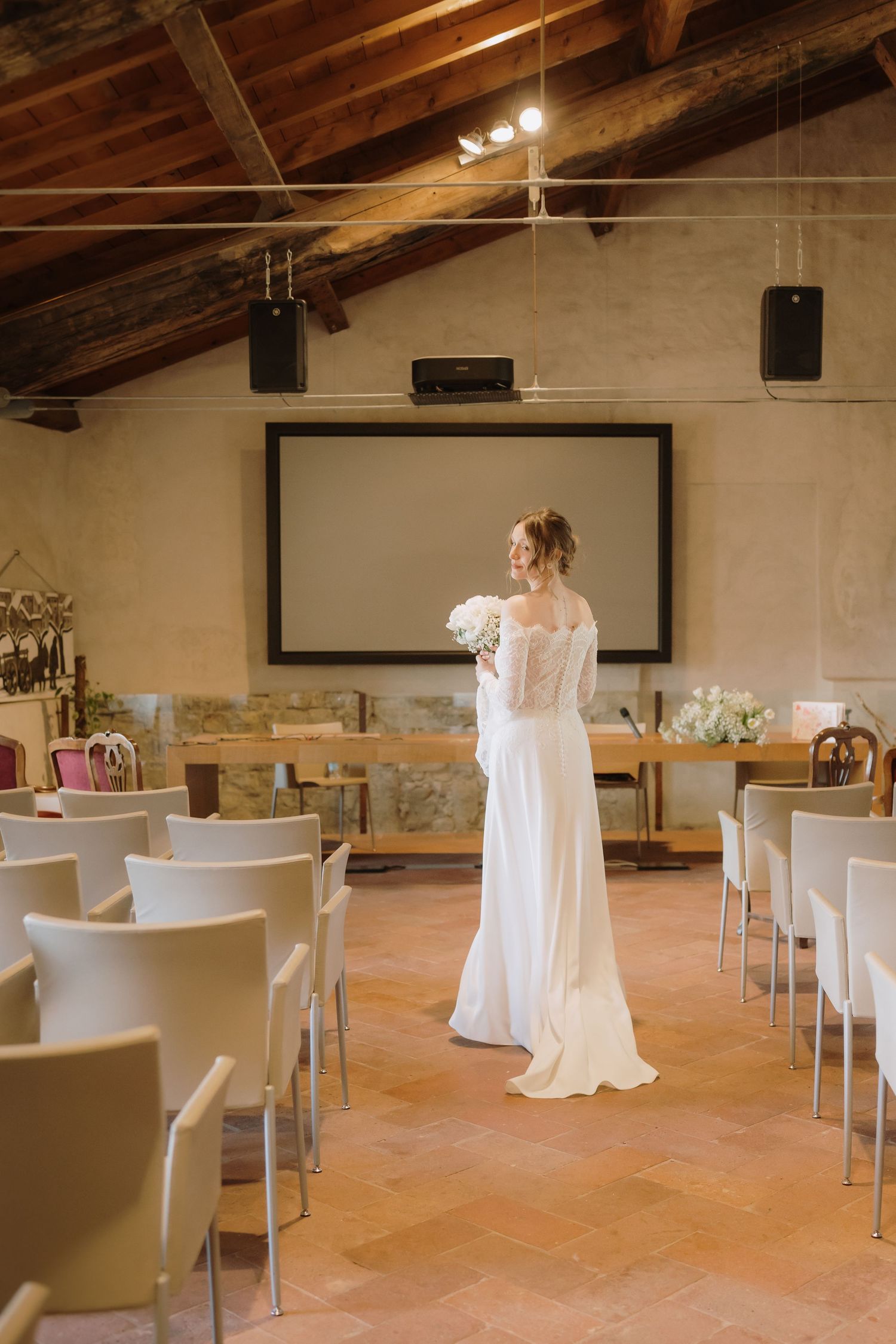 Bride in off-shoulder white gown stands alone in rustic ceremony hall with terracotta floors.