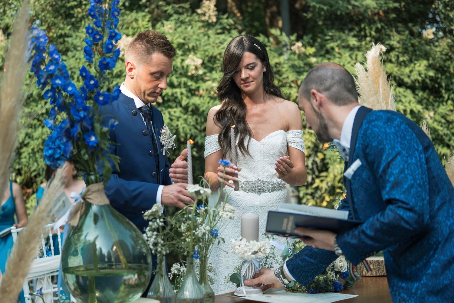 Couple signing wedding documents outdoors with officiant, blue floral arrangements, white dress and navy suit.