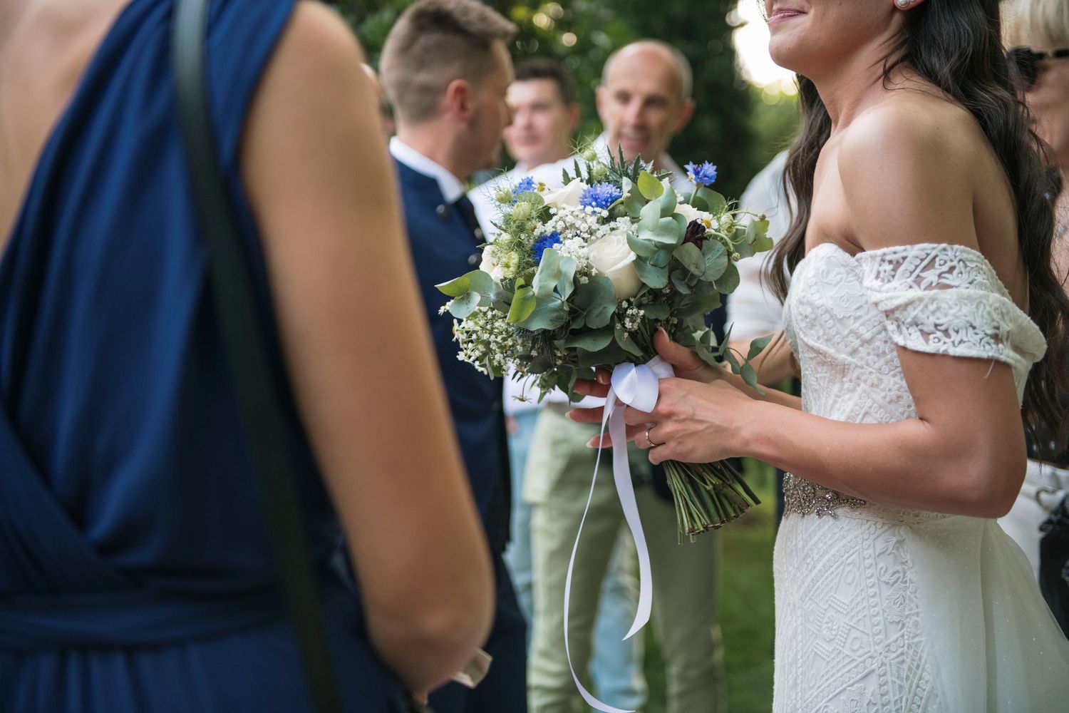 Bride in lace off-shoulder gown holds white and blue bouquet while mingling with wedding guests.