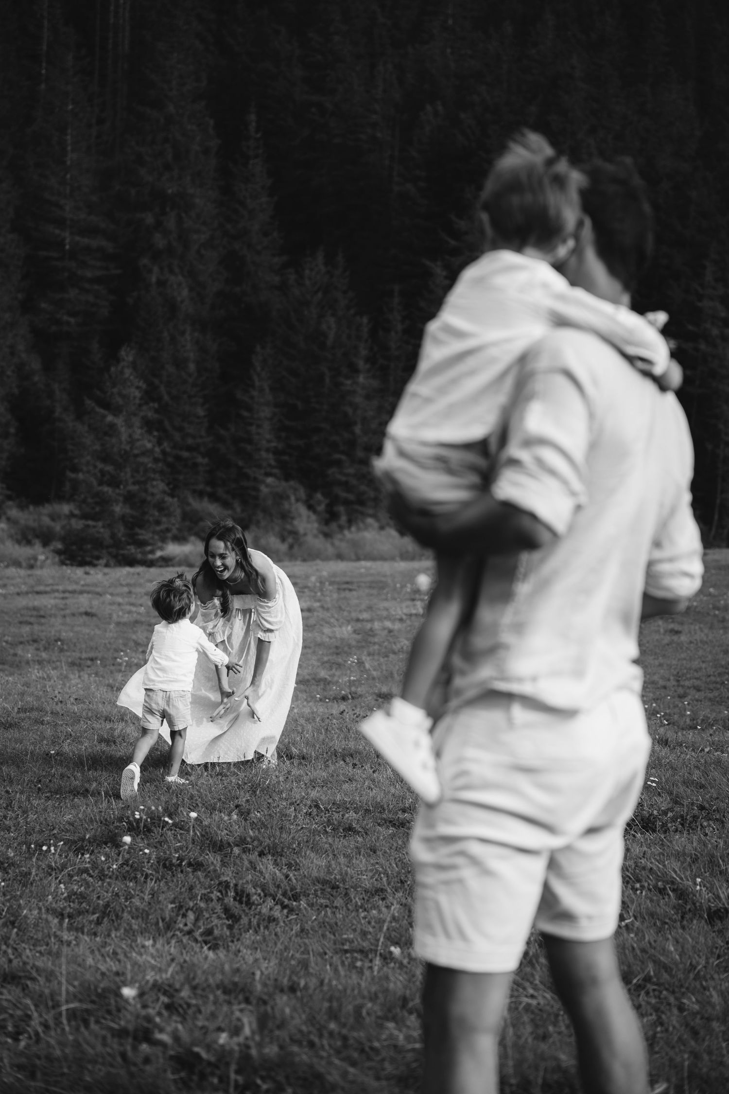 Black and white photo of a mother kneeling to greet her toddler on a grassy field with a man holding a child nearby.
