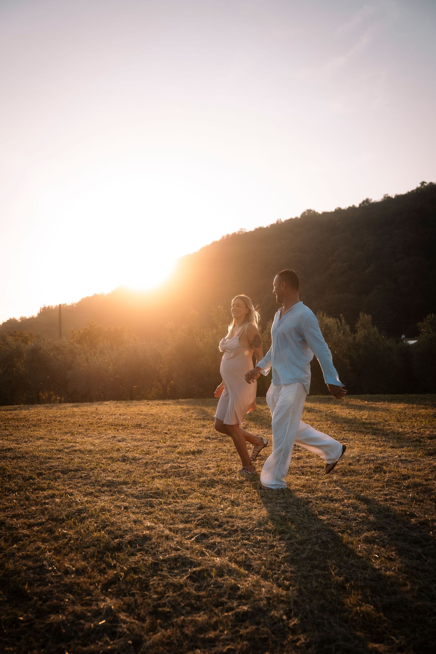 A joyful couple runs hand in hand across a sunlit field with a glowing mountain sunset behind them.
