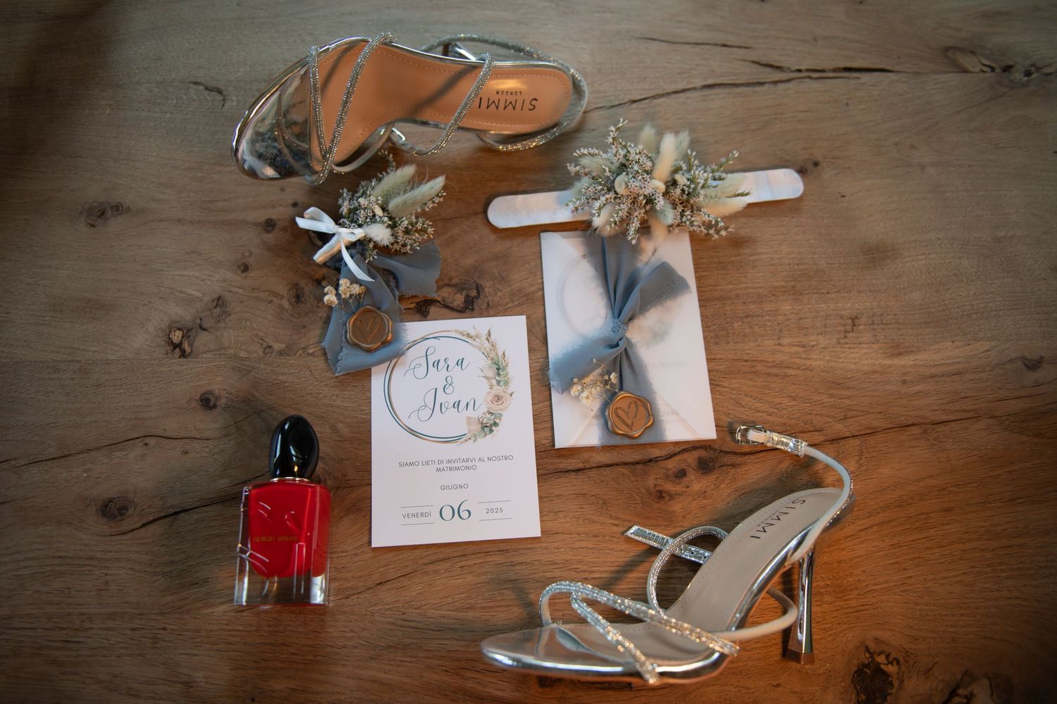 Silver wedding heels, floral garter, perfume, and elegant invitation flat lay on rustic wood.