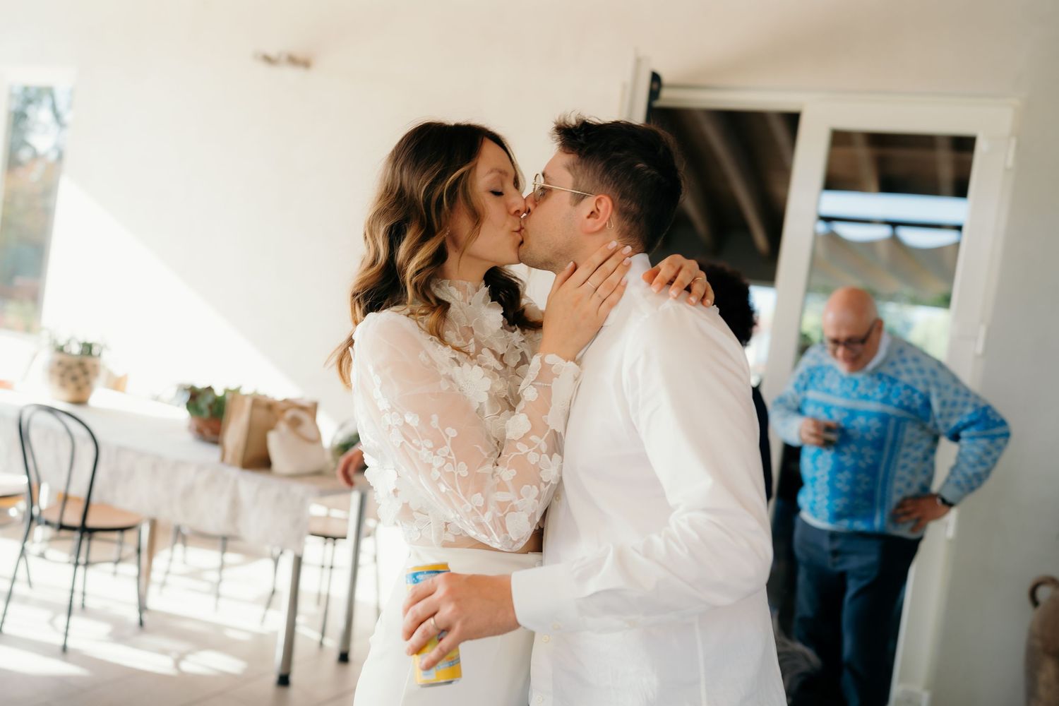 Newlyweds share romantic kiss at bright indoor wedding reception while guest stands nearby.