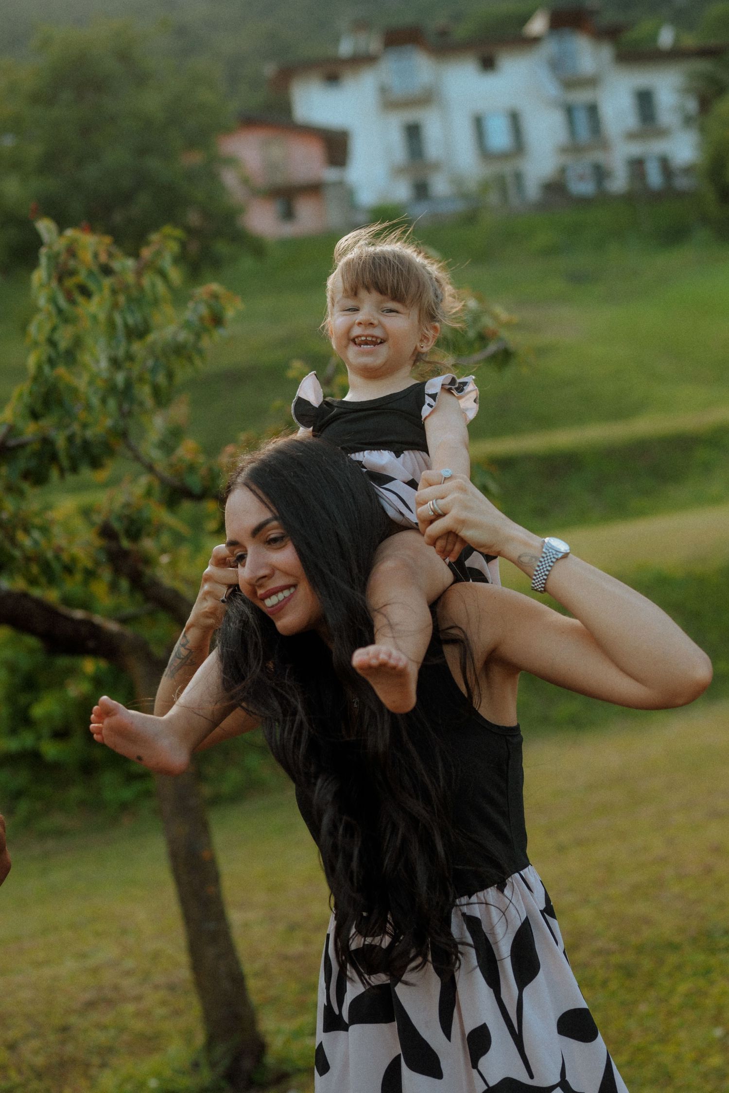 Una madre sorridente porta sulle spalle il suo bambino che ride, in un giardino verdeggiante vicino a una casa bianca.