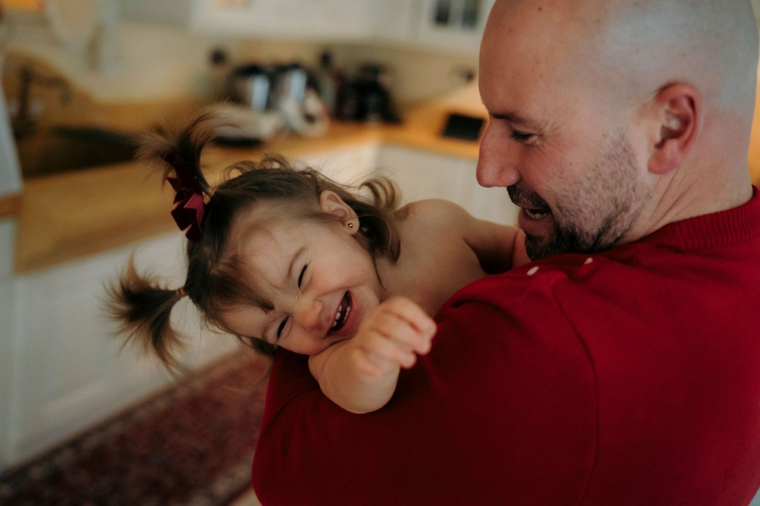 A bald father in a red shirt laughs while holding his giggling young daughter in a cozy kitchen.