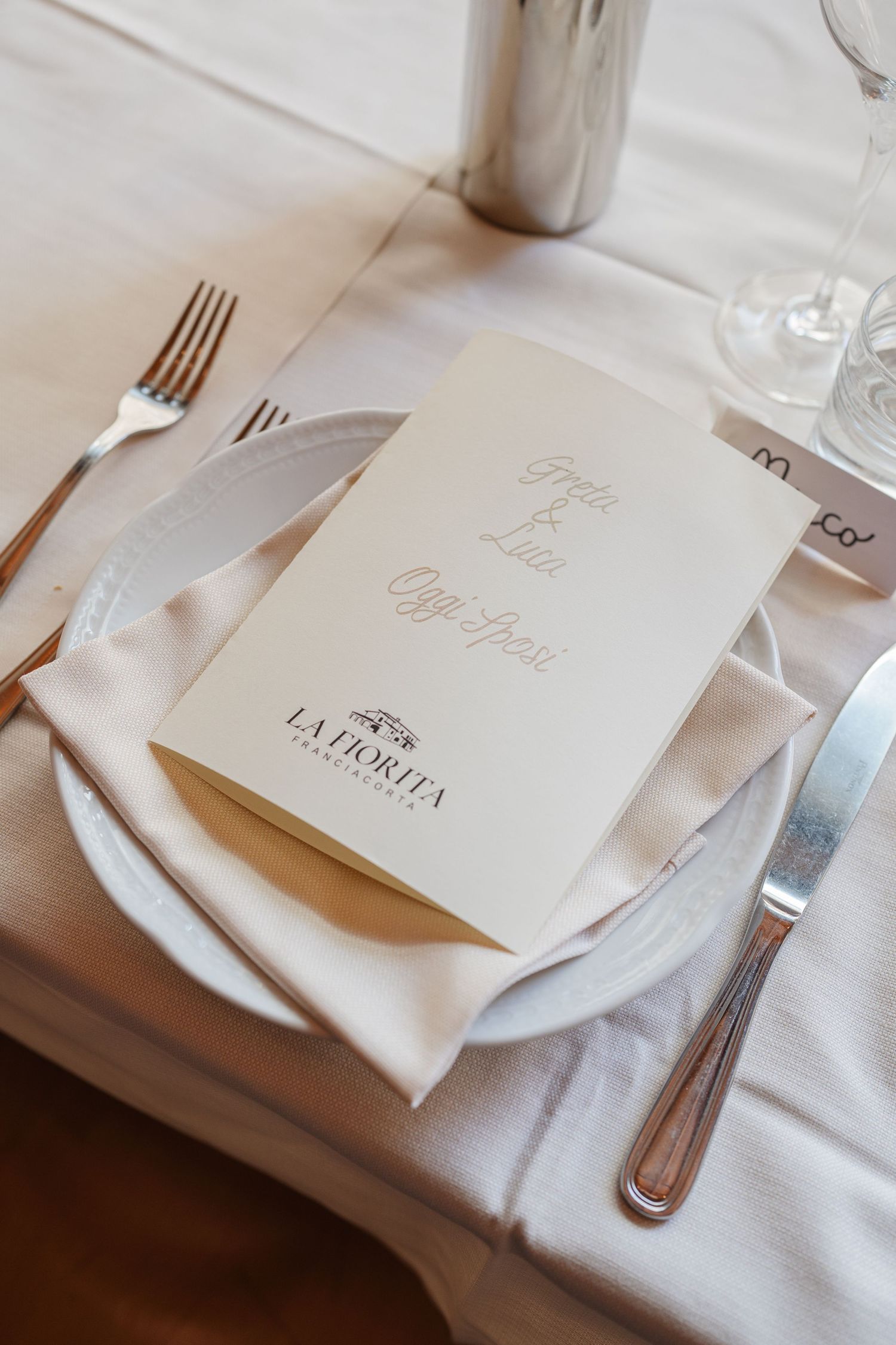Elegante mise en place nuziale presso La Fiorita, con menu color avorio, tovagliolo di lino, posate d'argento e bicchieri su tovaglia bianca