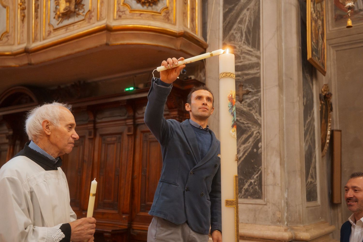Young man lighting large ceremonial candle assisted by priest during church baptism service.