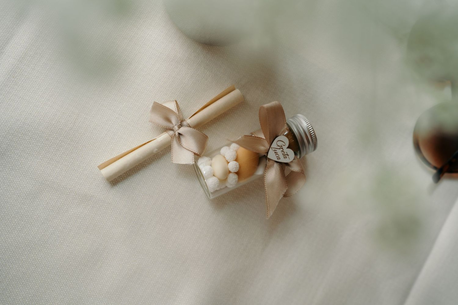 Eleganti bomboniere con nastri in raso beige, ciondoli a forma di cuore e confetti su uno sfondo di morbido tessuto bianco.