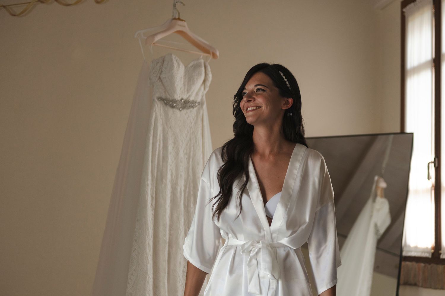 Smiling bride in white satin robe admiring her wedding dress hanging on display in a bright elegant room.