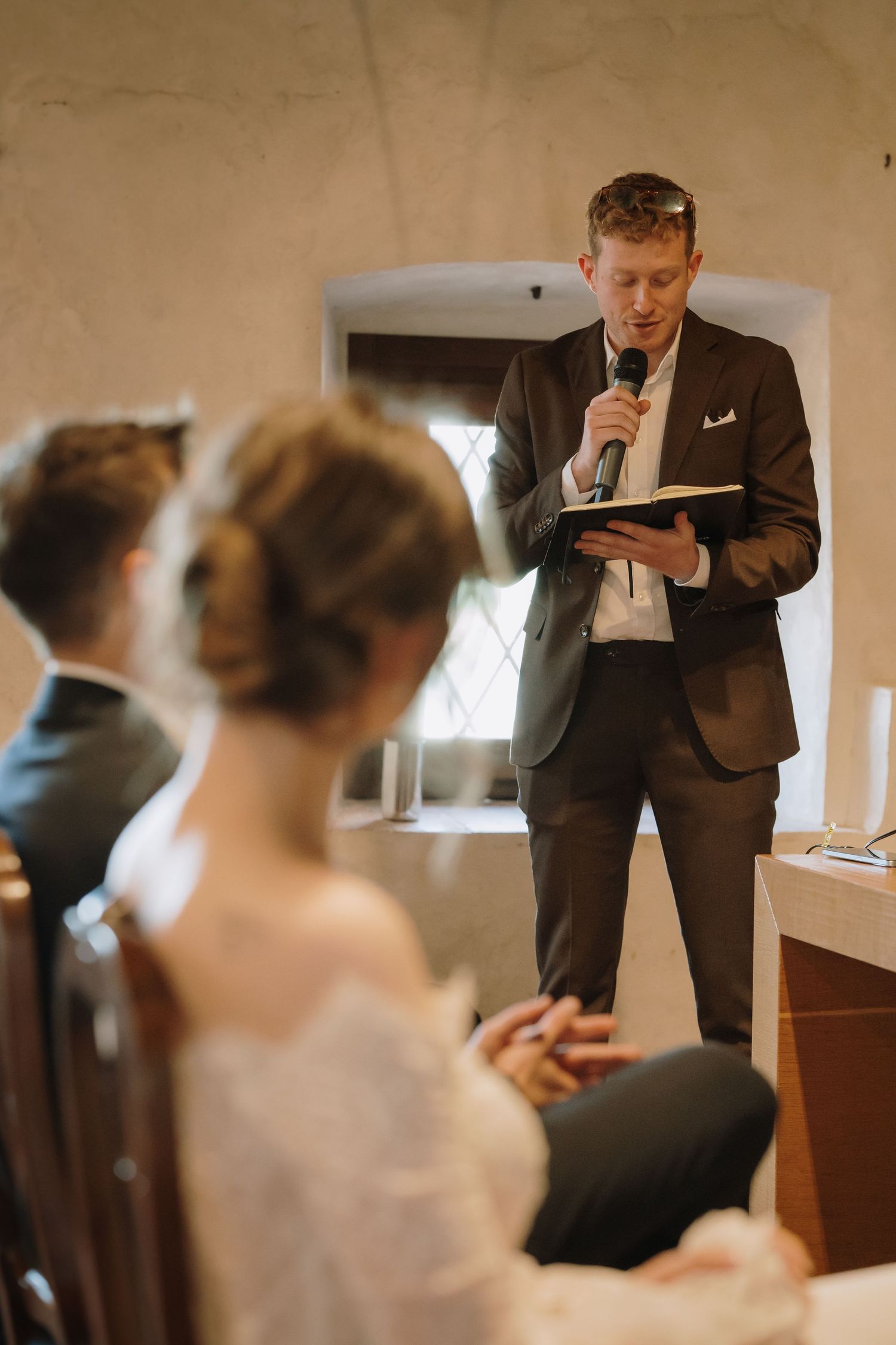 A man in a dark suit reads from a book into a microphone at a wedding ceremony while the bride and groom listen.