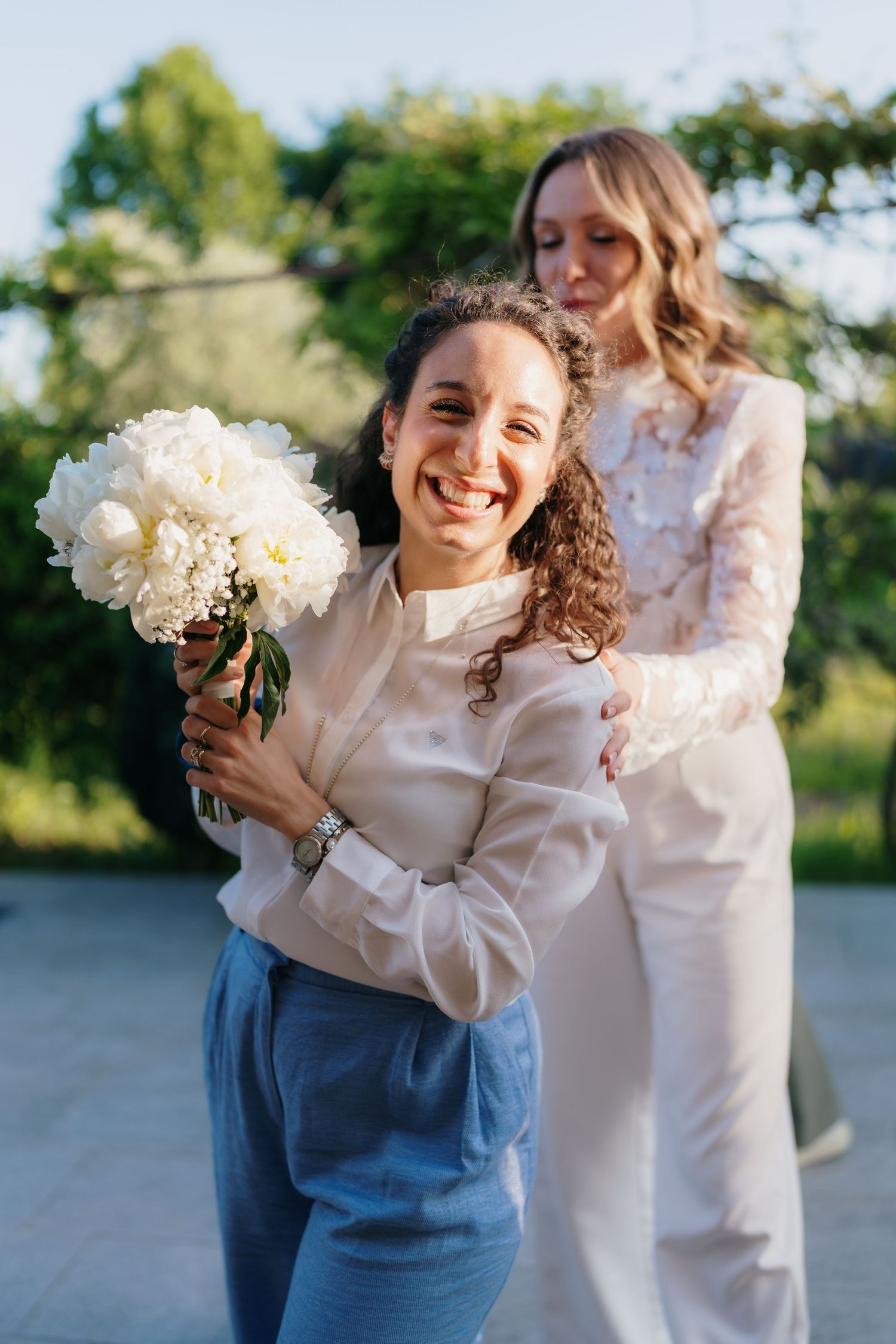 Two women in white outfits celebrate outdoors, one smiling holding a white peony bouquet while the other embraces her from behind.
