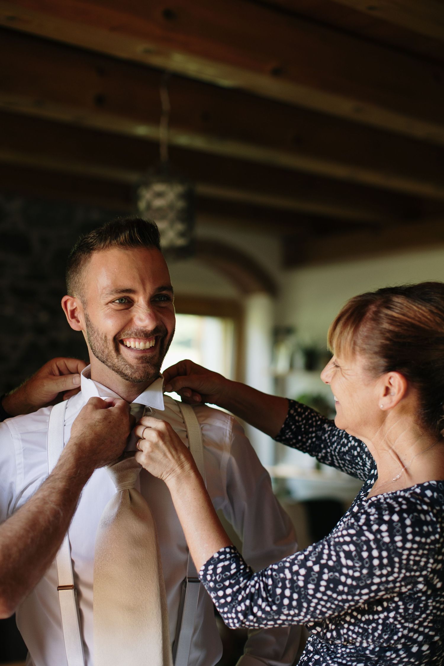 Smiling groom getting his tie adjusted by a woman in a bright rustic room before his wedding ceremony.
