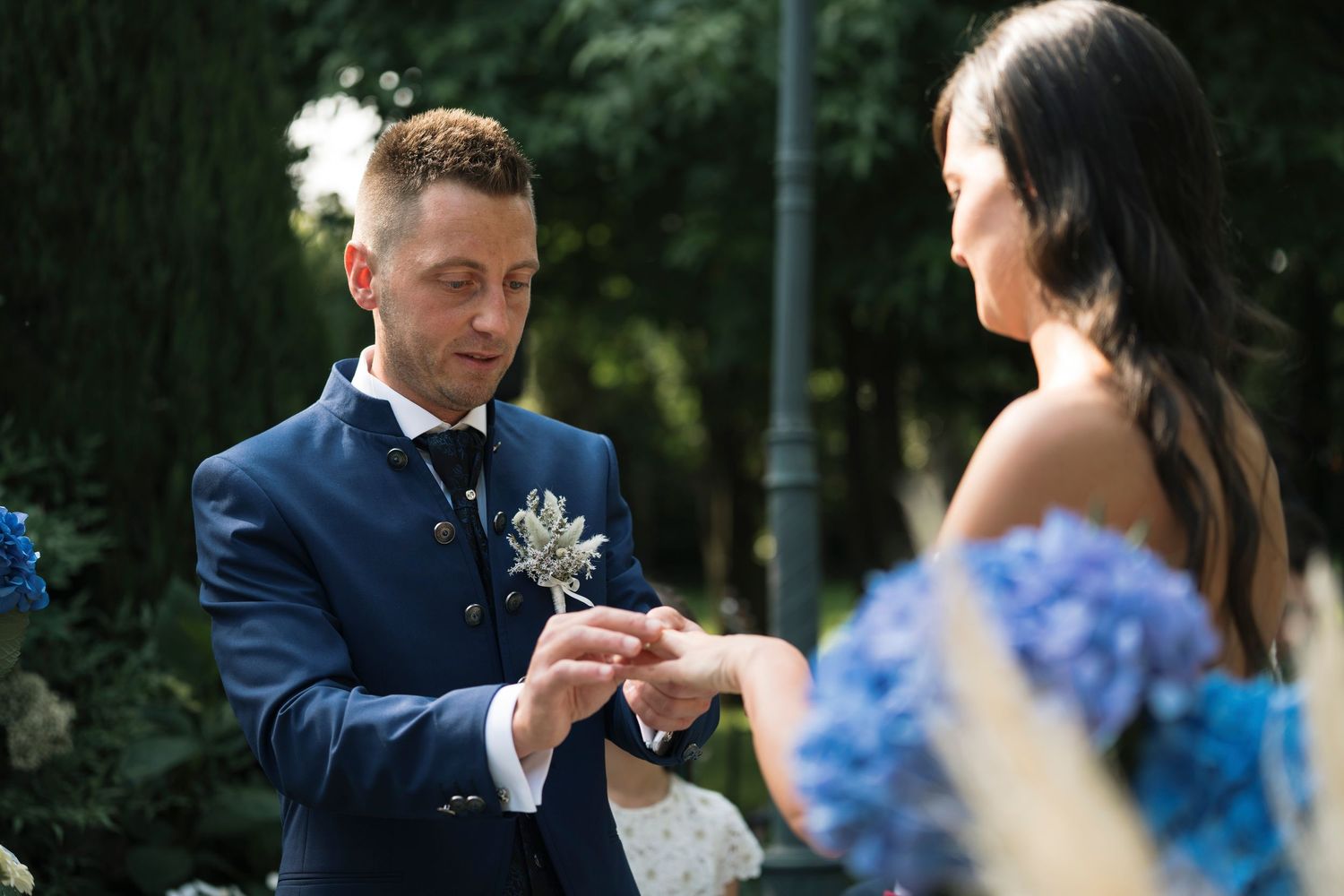 Groom in navy suit places ring on brides finger during outdoor wedding ceremony.