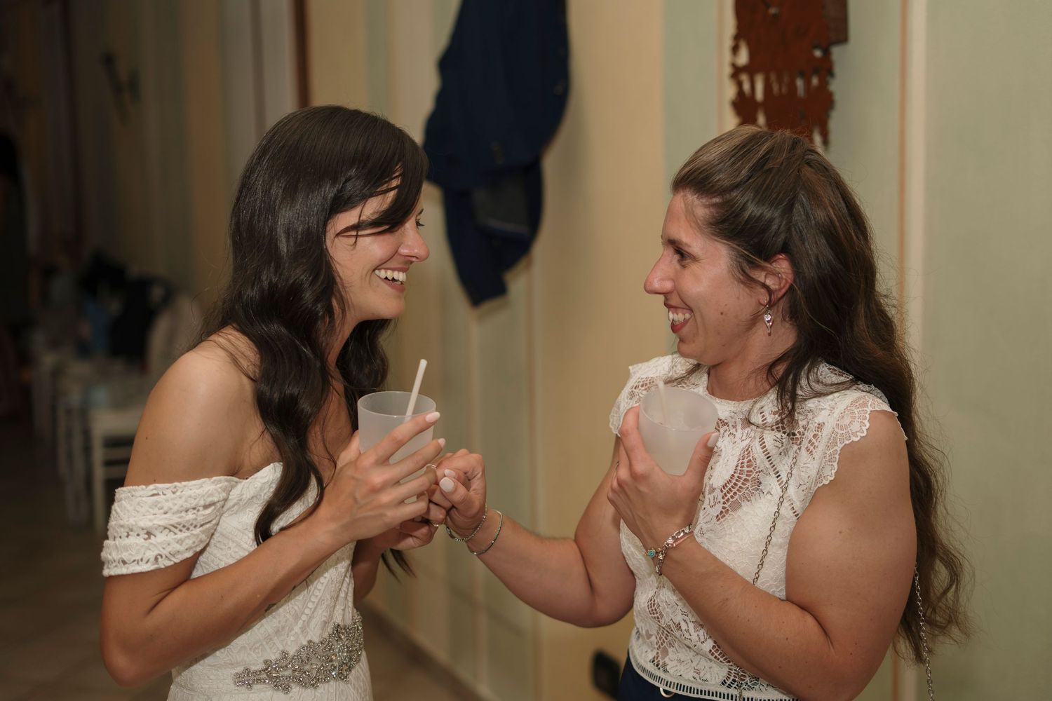 Two women in white dresses laughing and sharing drinks together at a wedding celebration.