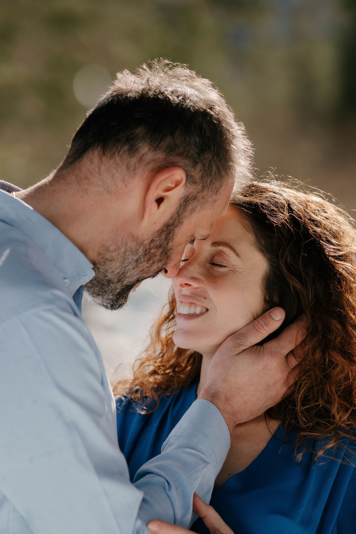 Tender close-up of bearded man kissing curly-haired woman on forehead outdoors.