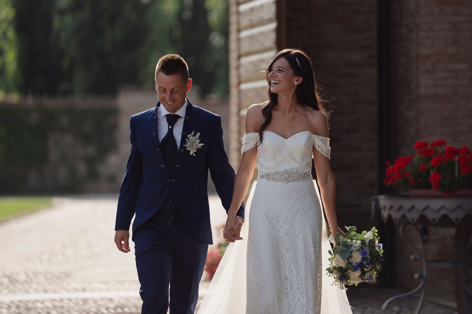 Newlyweds walk hand in hand, groom in navy suit and bride in lace gown at wedding venue.