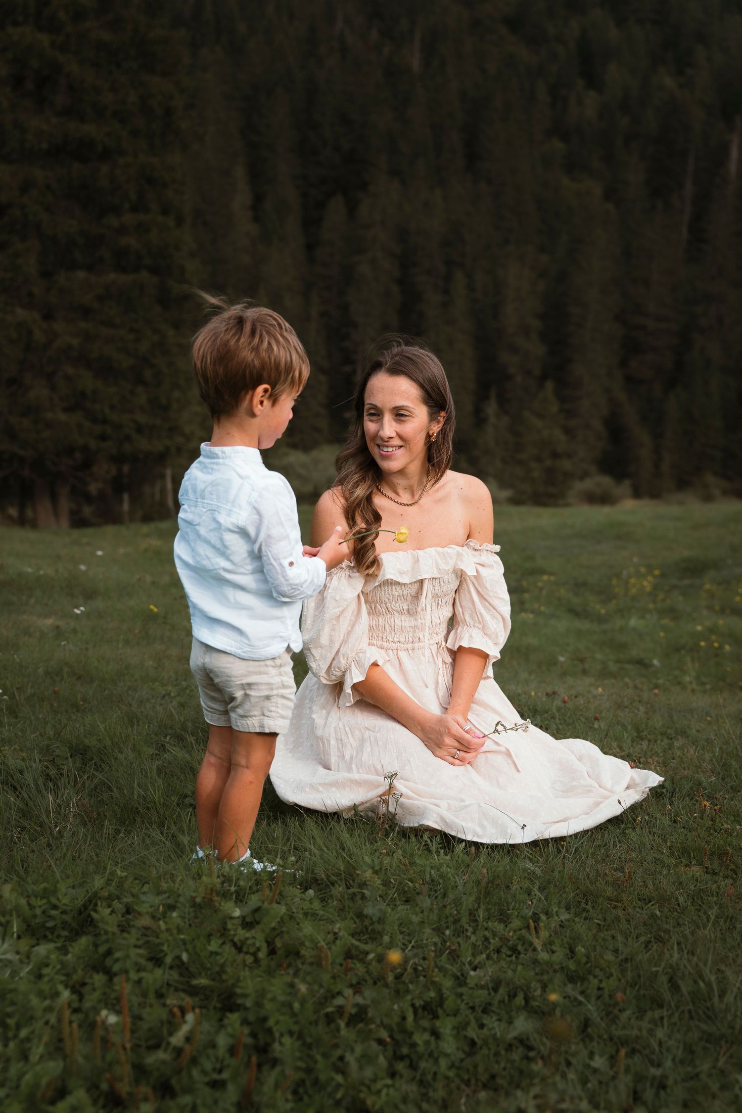 A woman in a white off-shoulder dress sits on grass smiling at her young son standing before her in nature.