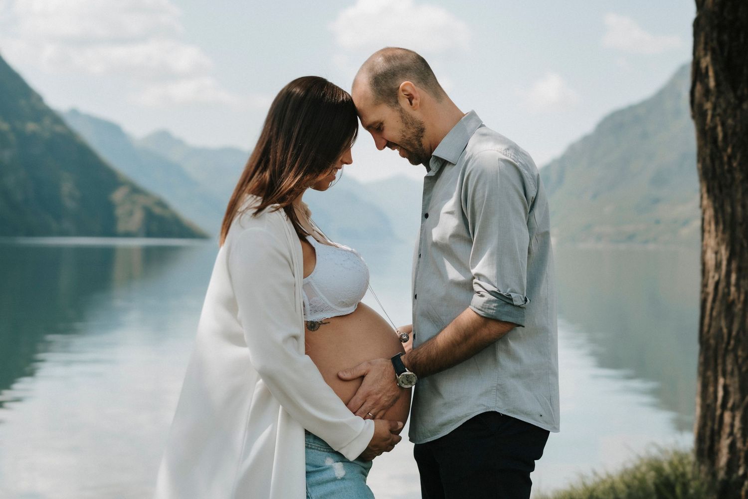 Expecting couple tenderly touching pregnant belly by a serene mountain lake on a clear day.