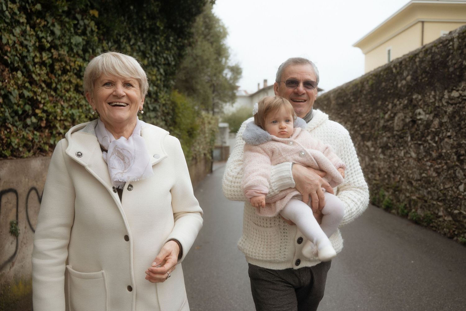 Smiling grandparents walking outdoors on a tree-lined path, grandfather carrying a baby dressed in pink.