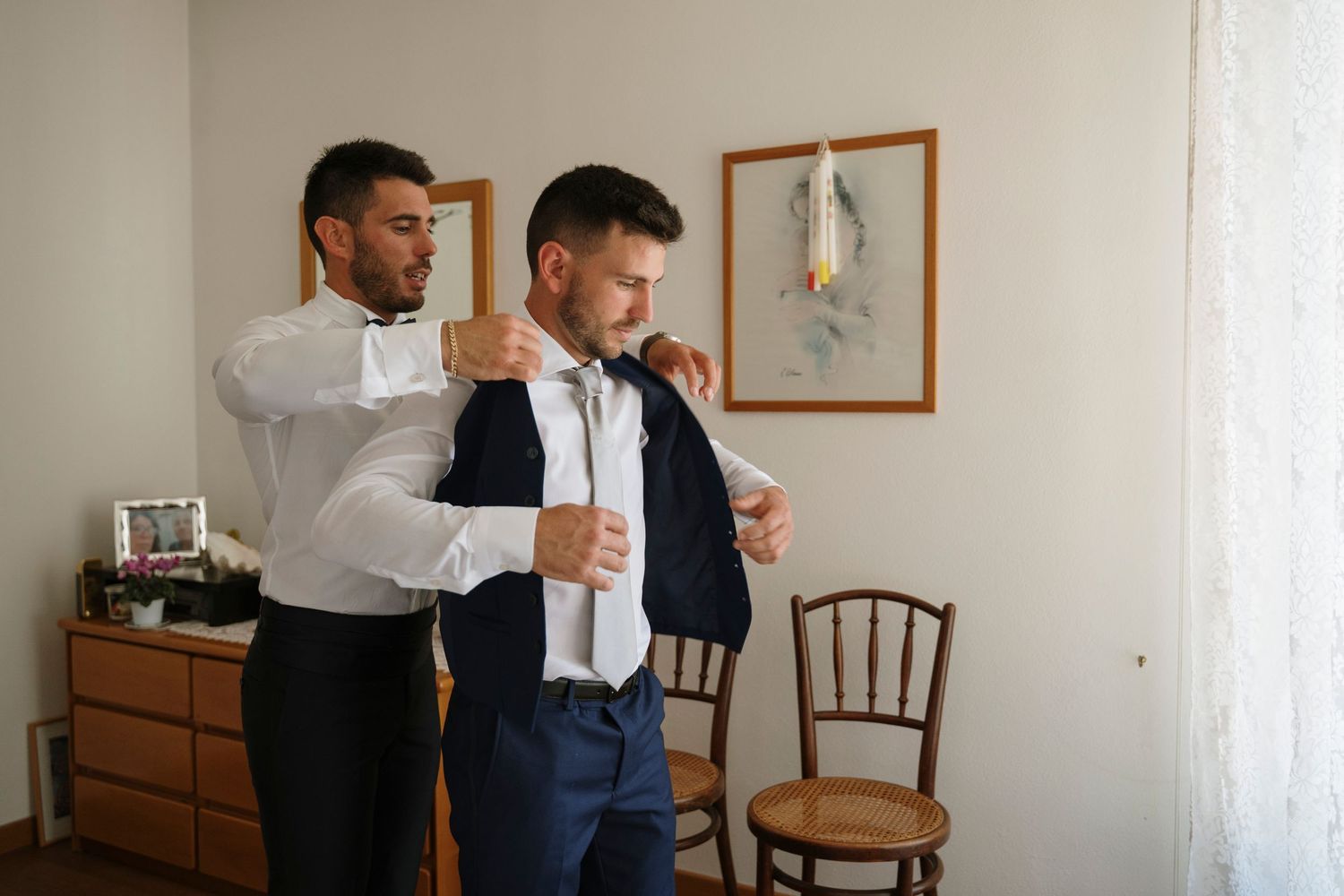Groomsman helping groom adjust his tie while getting dressed for a wedding ceremony indoors.