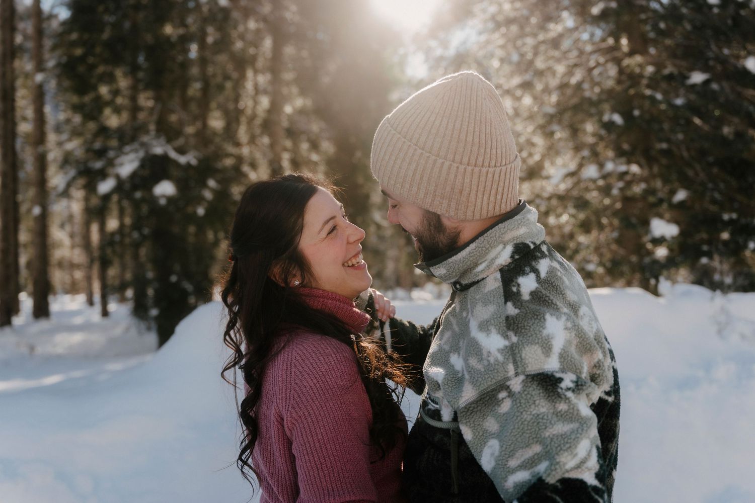 Happy couple embracing and laughing together in a sunny snowy forest setting.