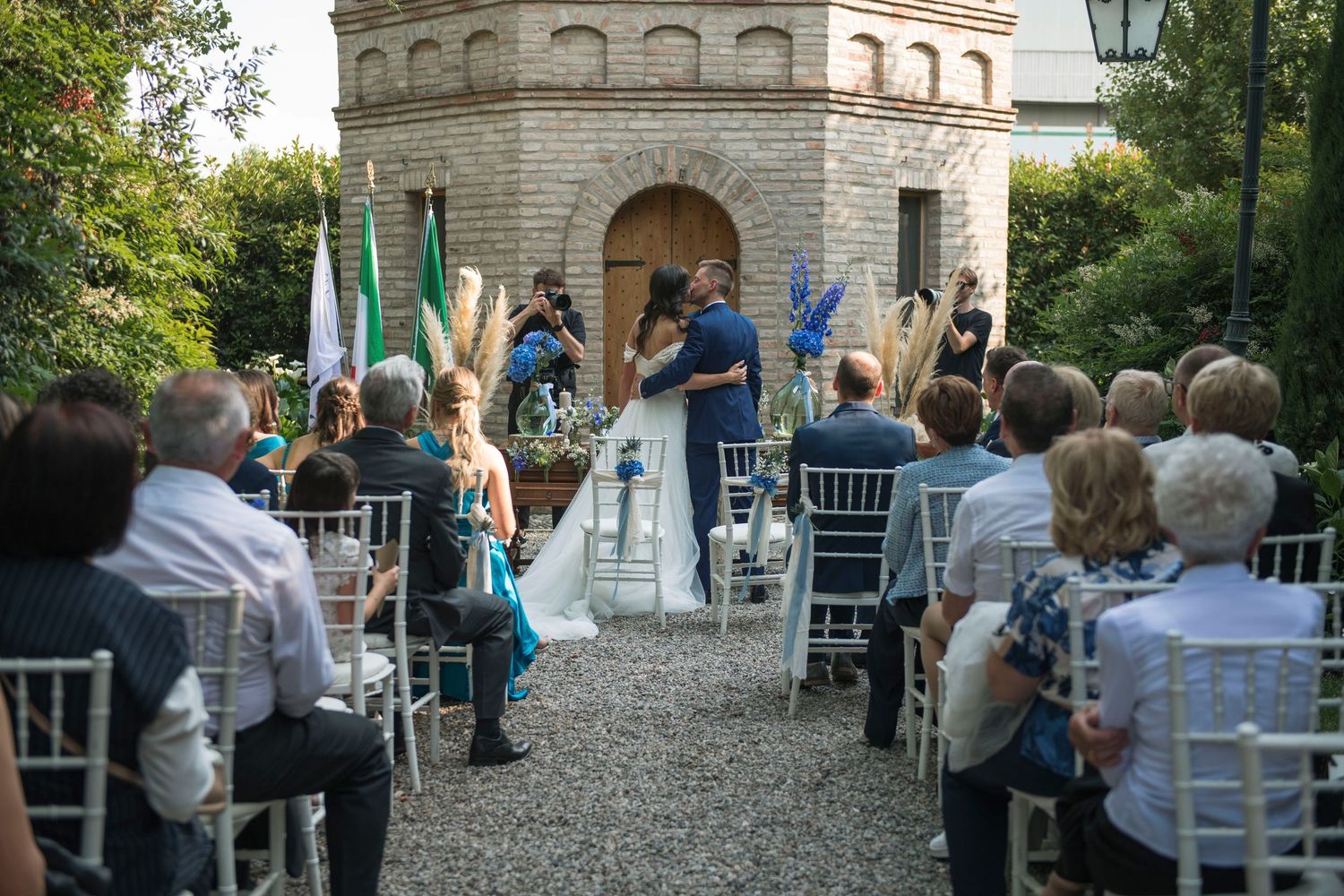 Outdoor wedding ceremony with guests seated as couple kisses at the altar near a stone tower.
