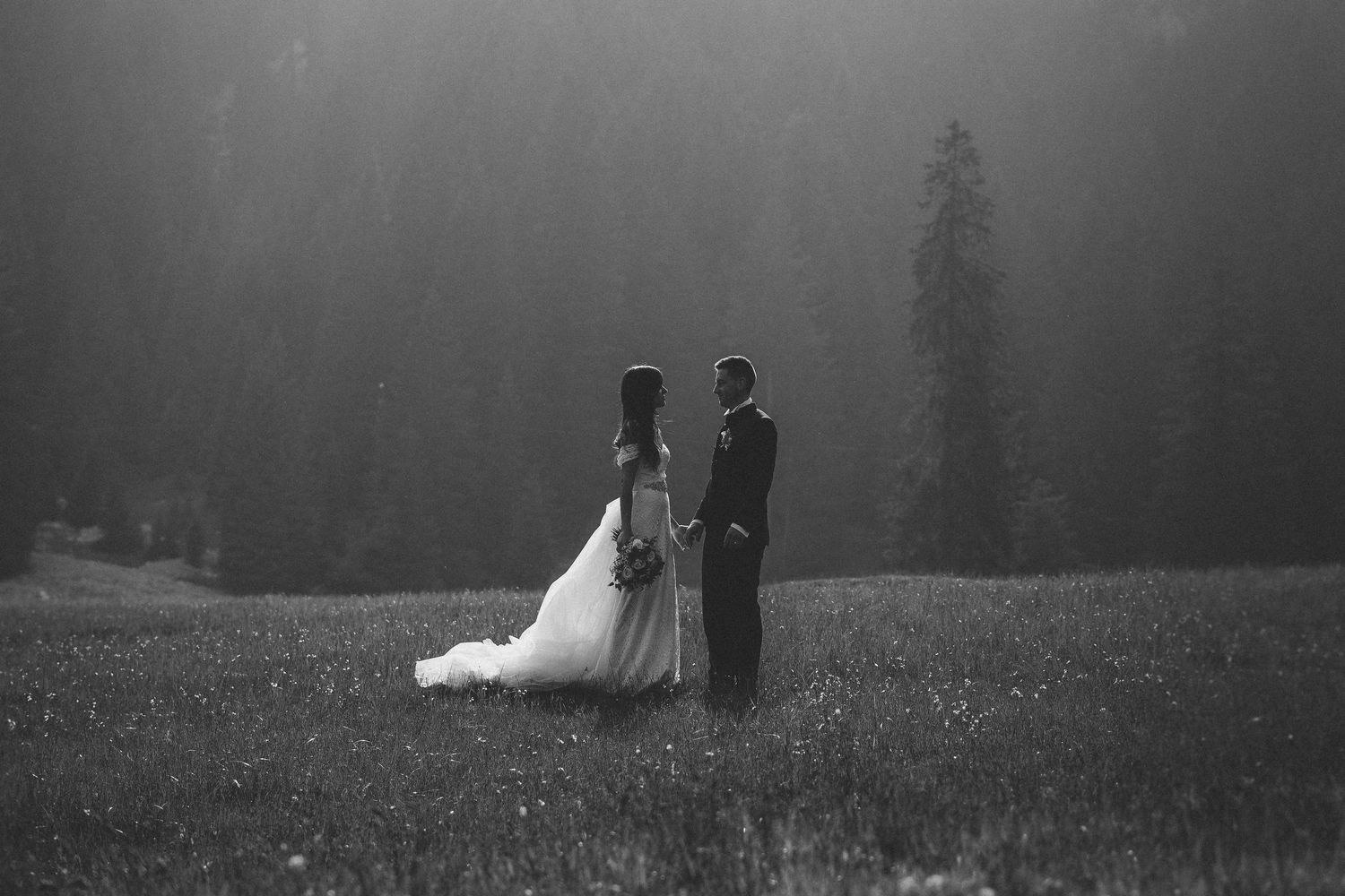 Black and white photo of a bride and groom standing in a misty meadow with tall evergreen trees in the background.