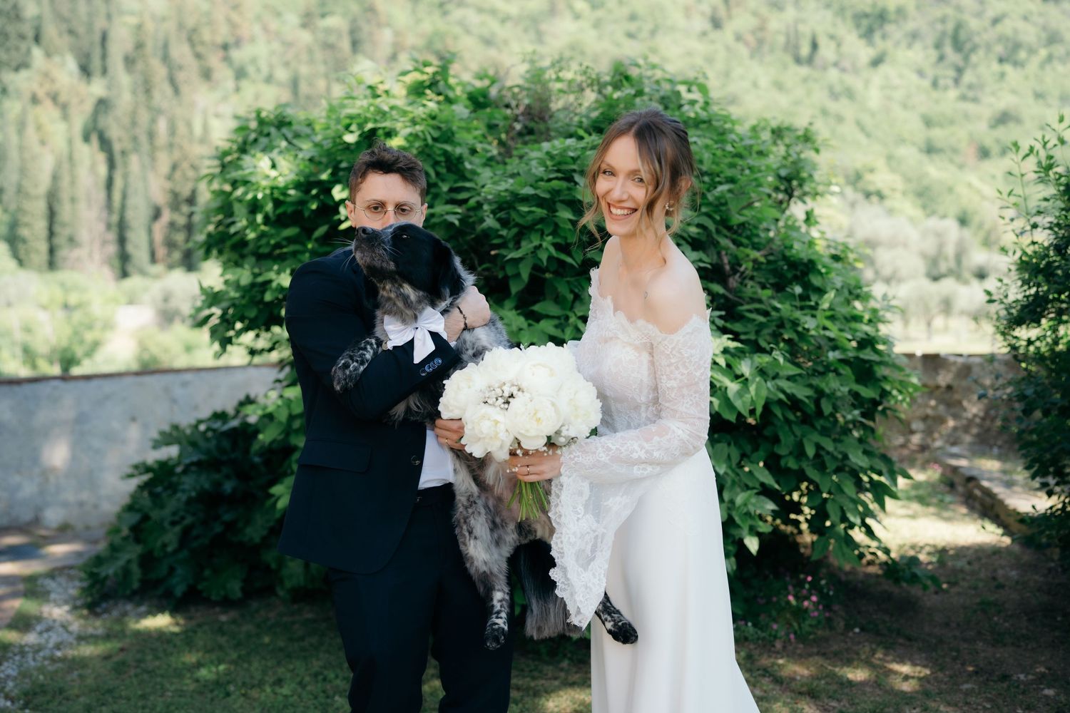 Bride and groom pose outdoors holding a dog and white bouquet on their wedding day.