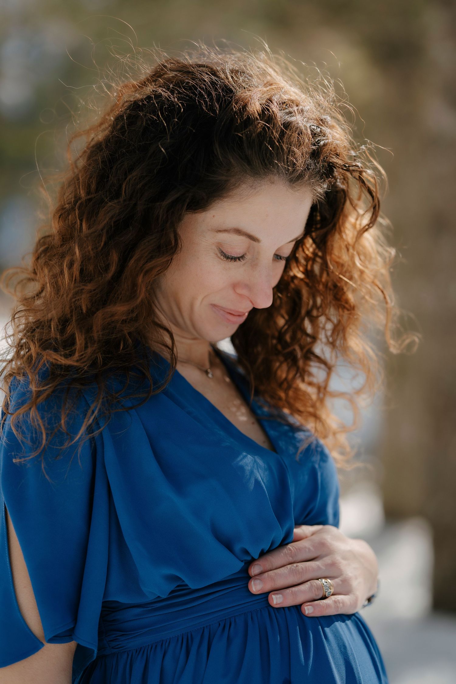 Curly-haired pregnant woman in blue dress gently holding her baby bump outdoors.
