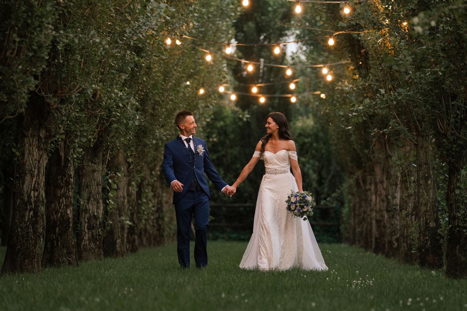 Bride and groom walk hand-in-hand through a tree-lined path adorned with string lights at dusk.