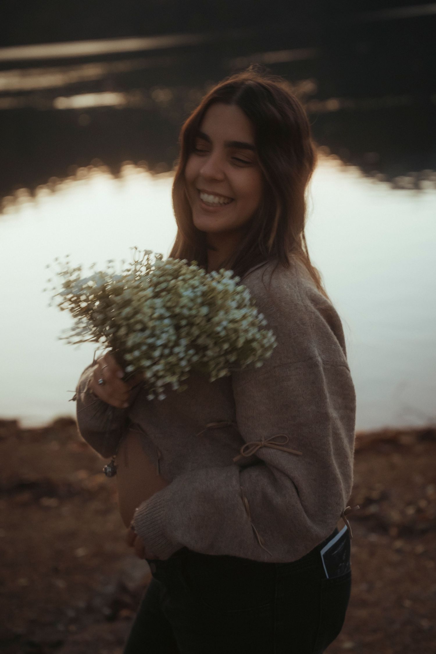 Smiling woman holding white wildflower bouquet beside reflective lake at dusk.