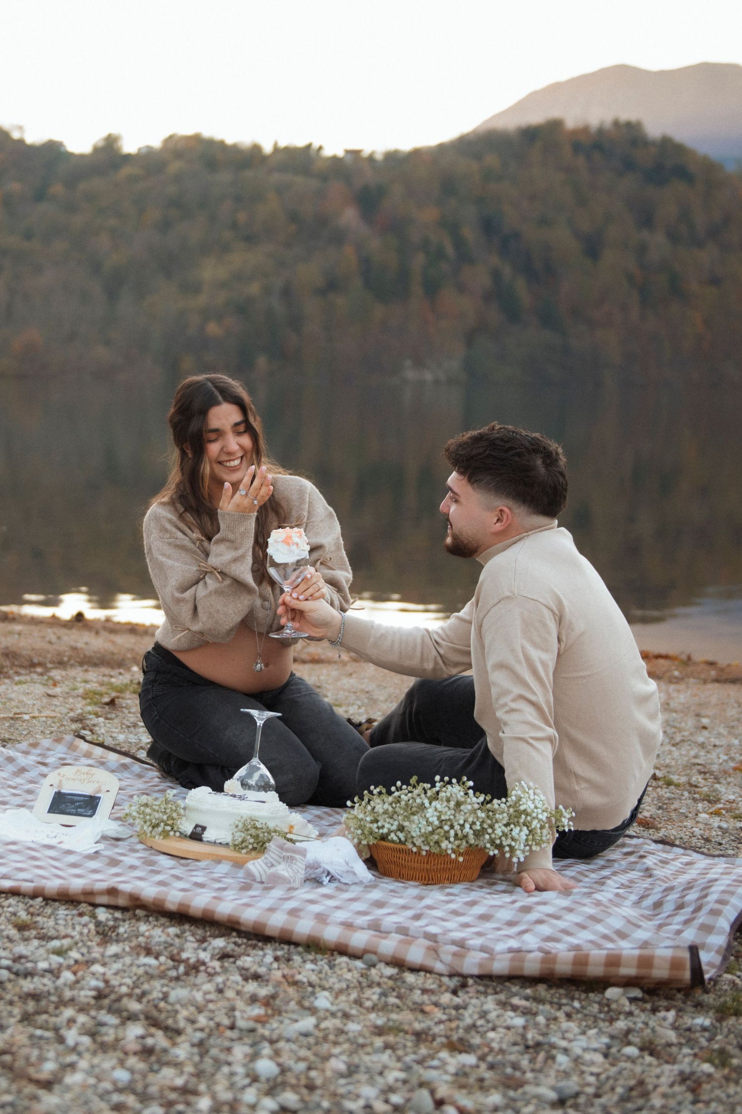 Pregnant couple enjoying romantic lakeside picnic with cake and flowers at sunset.