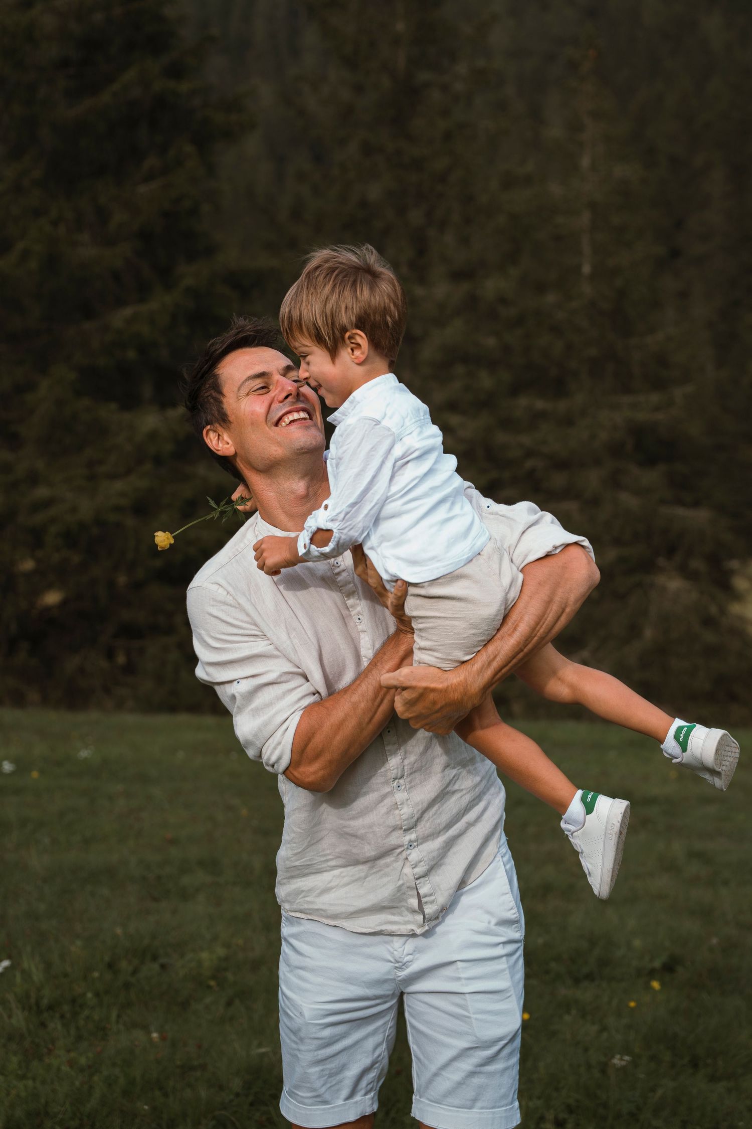A smiling father lifts his young son high in the air outdoors with a lush green forest backdrop.