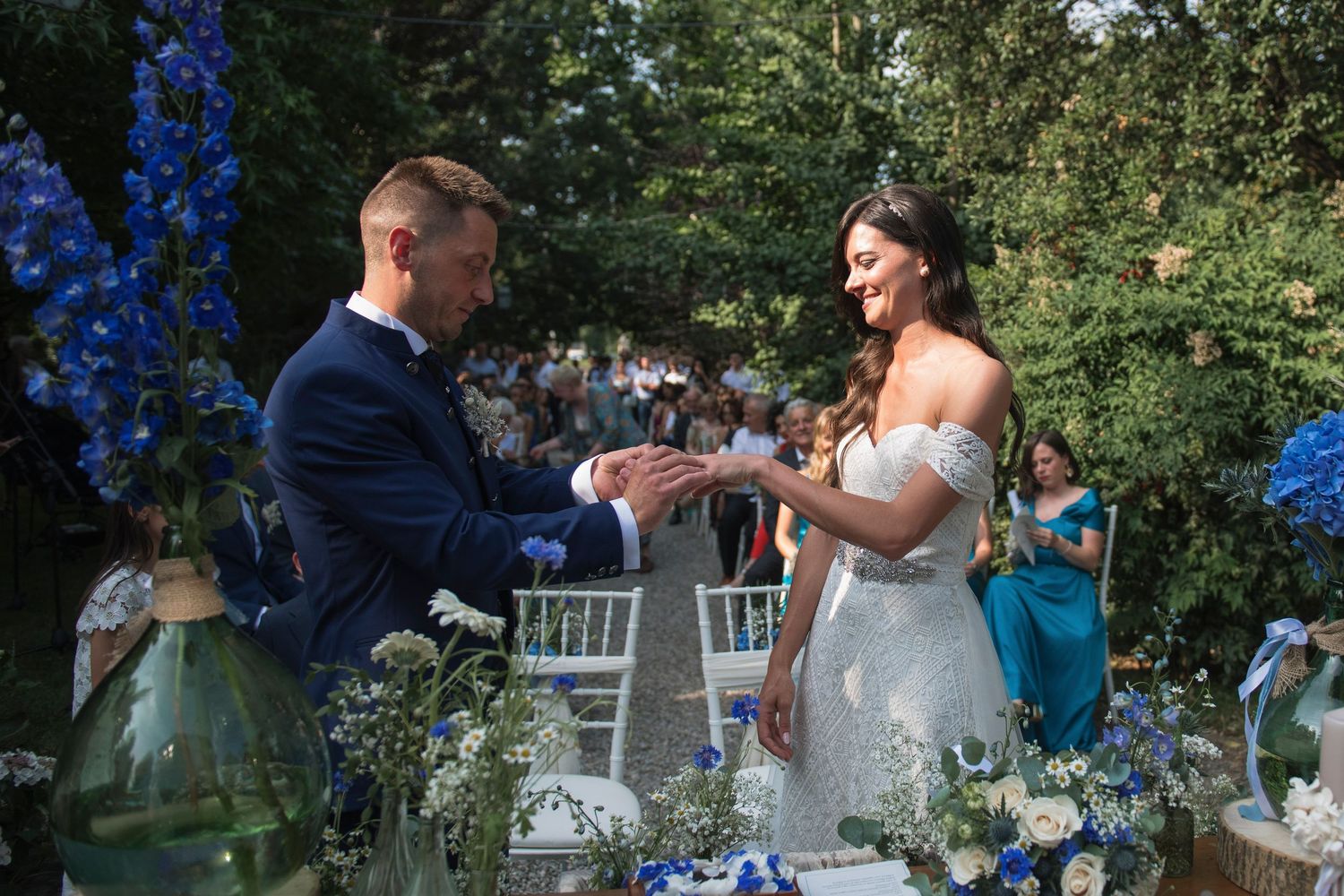 Groom placing ring on brides finger during outdoor wedding ceremony with blue floral decor.