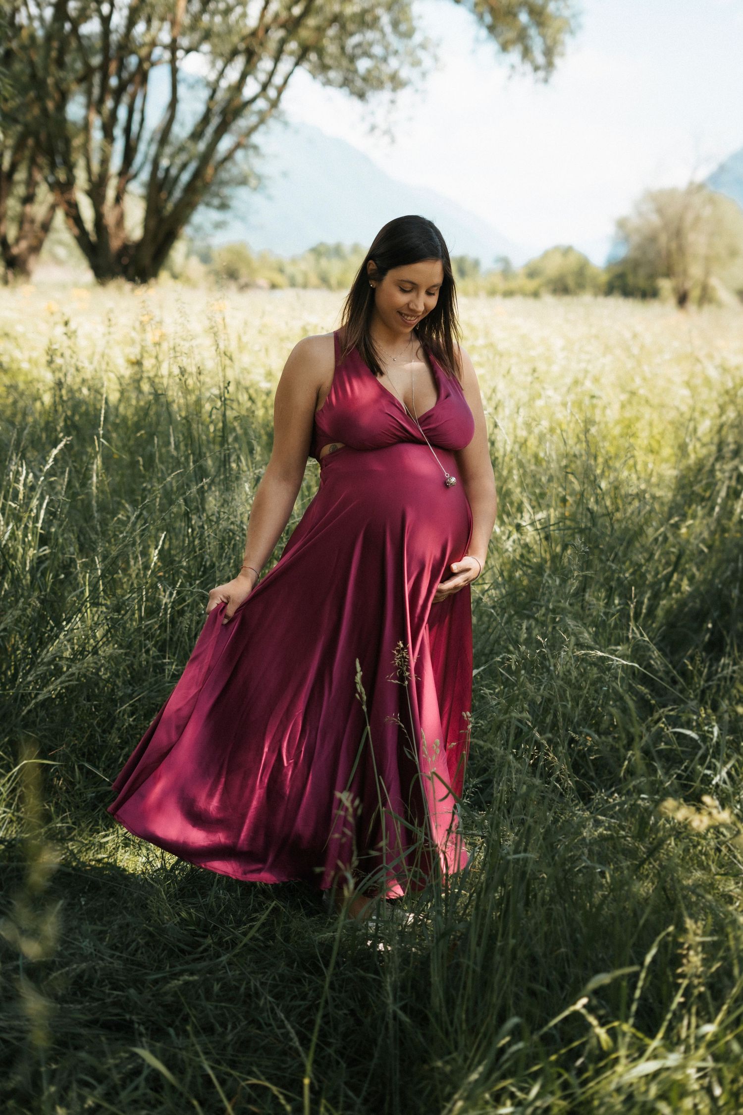 Pregnant woman in a flowing magenta dress standing in a lush green meadow with mountains in the background.