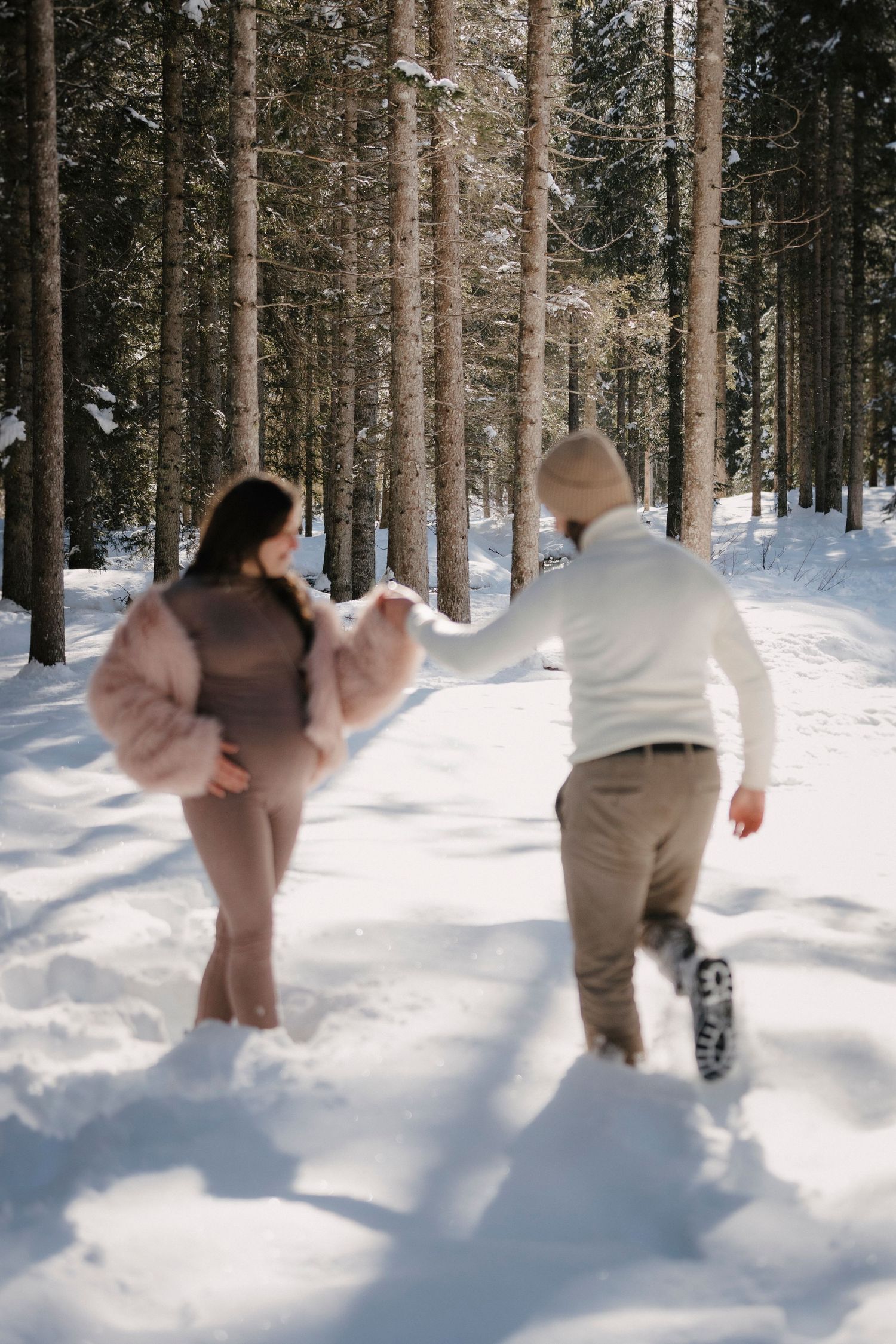 Pregnant couple holding hands and running playfully through a snow-covered pine forest in winter.