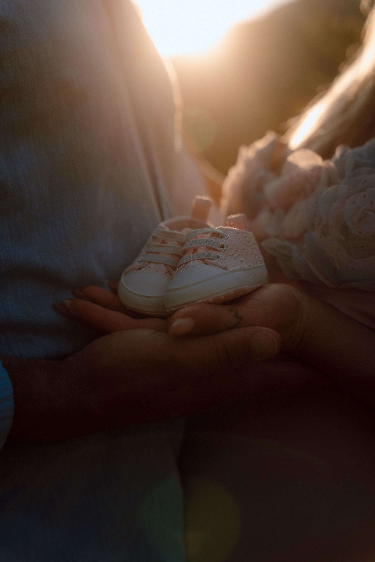 Couple holds tiny baby shoes together in soft golden sunlight, announcing their pregnancy.