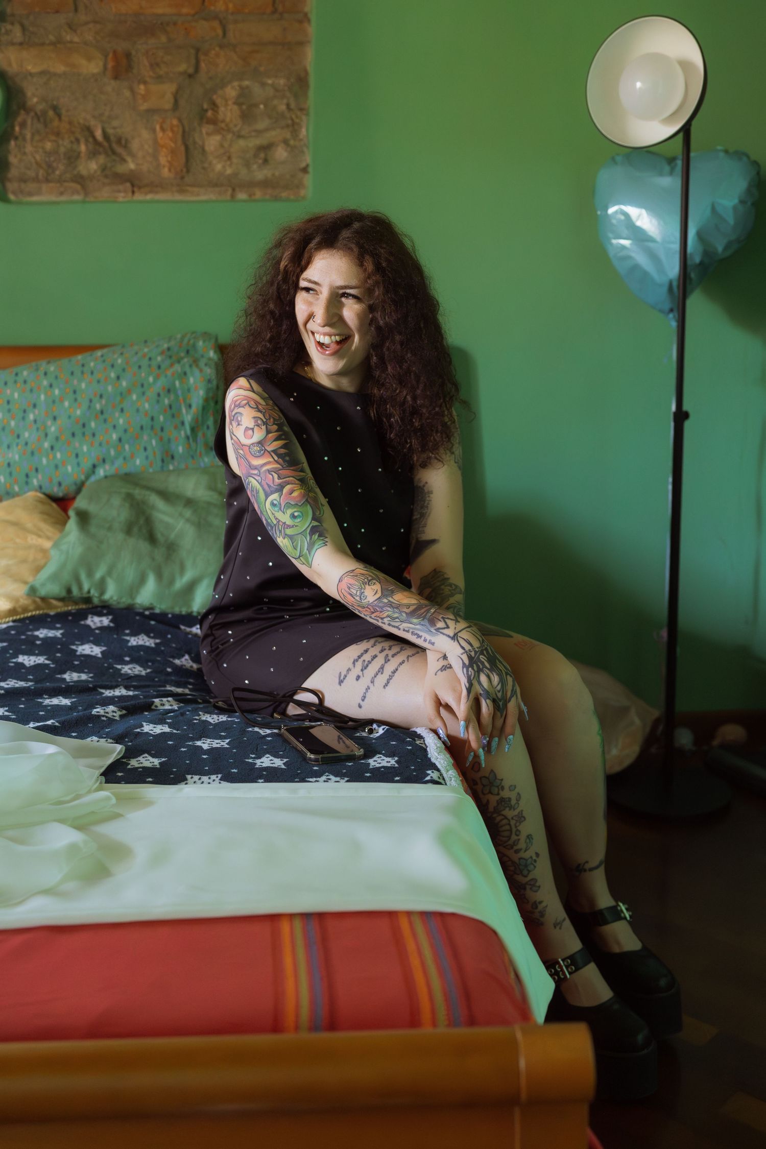 Smiling tattooed woman with curly hair sitting on a bed in a green room wearing a black dress and heels.