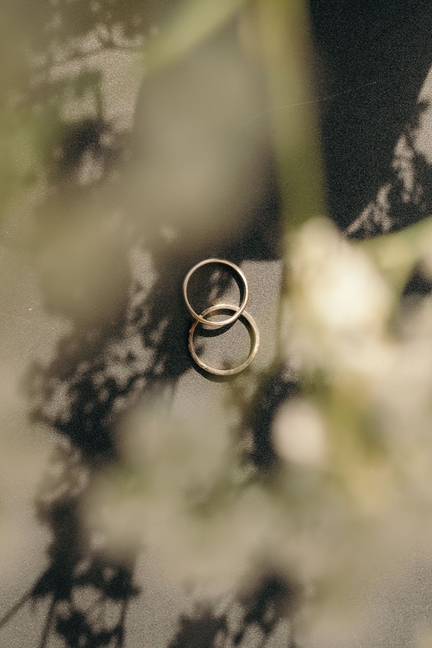 Two gold wedding bands rest together on a dark surface surrounded by soft floral bokeh.